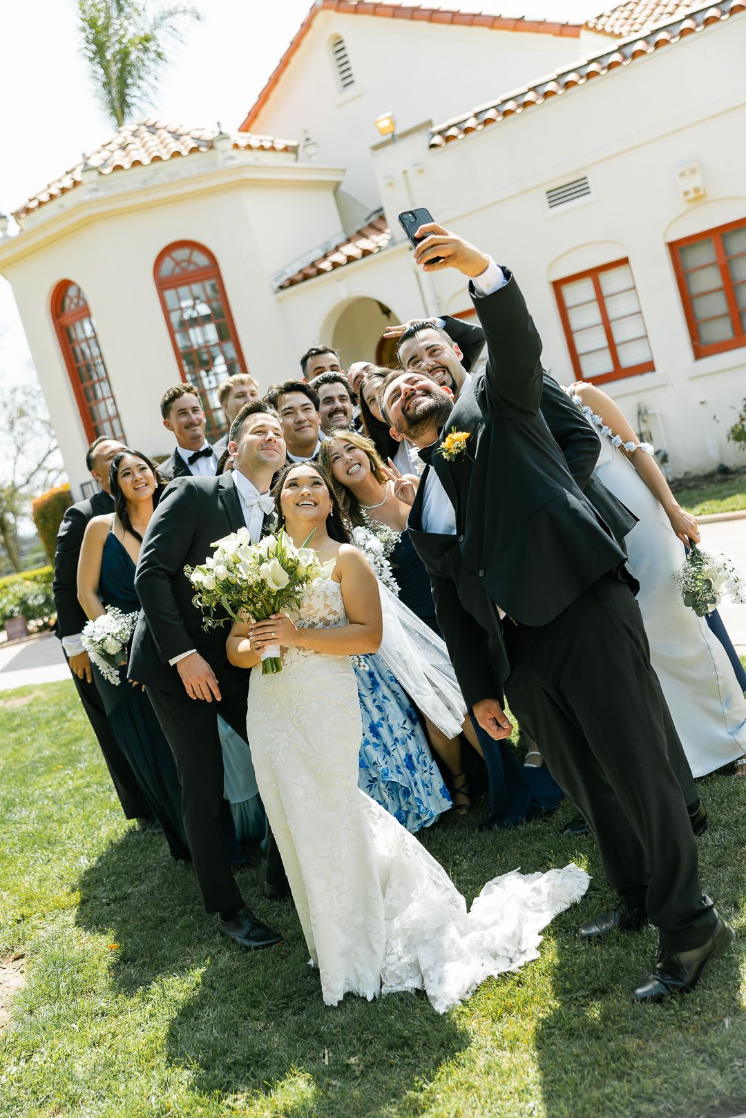 Wedding party poses together on sunny lawn outside Spanish-style building with white stucco walls and red tile roof.