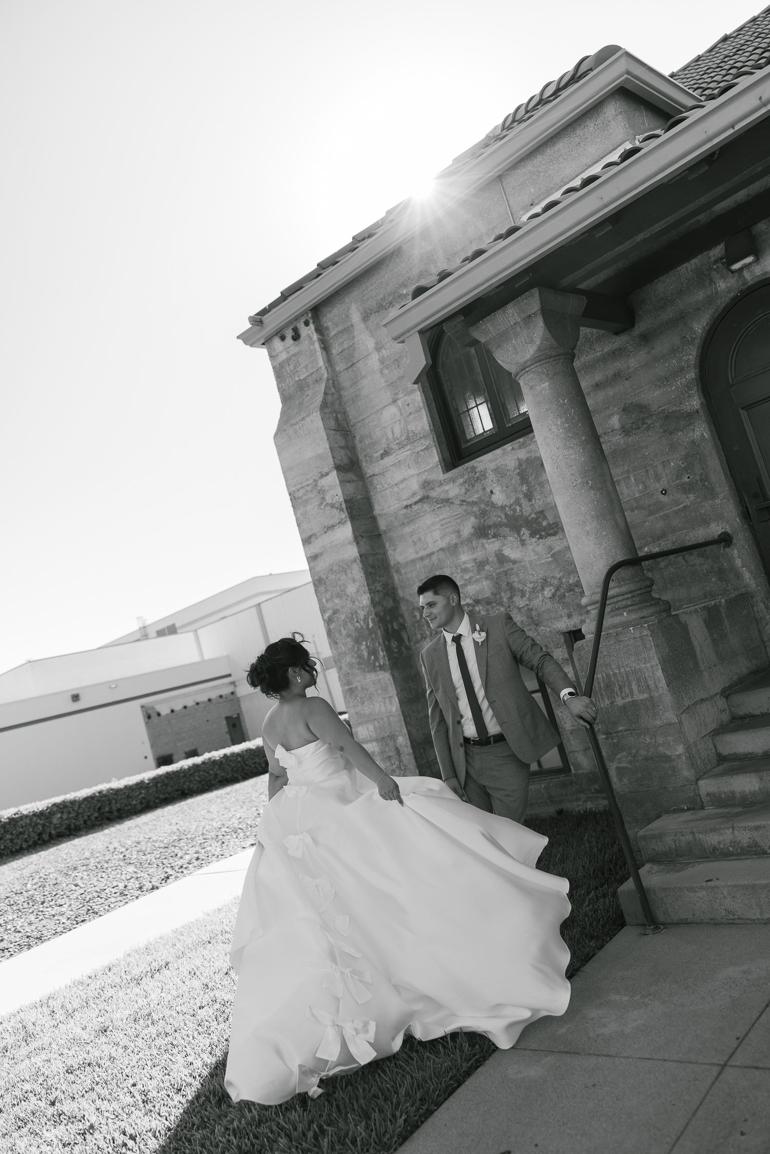 A wedding couple stands at the entrance of a stone building with sunlight peeking through on a bright sunny day.