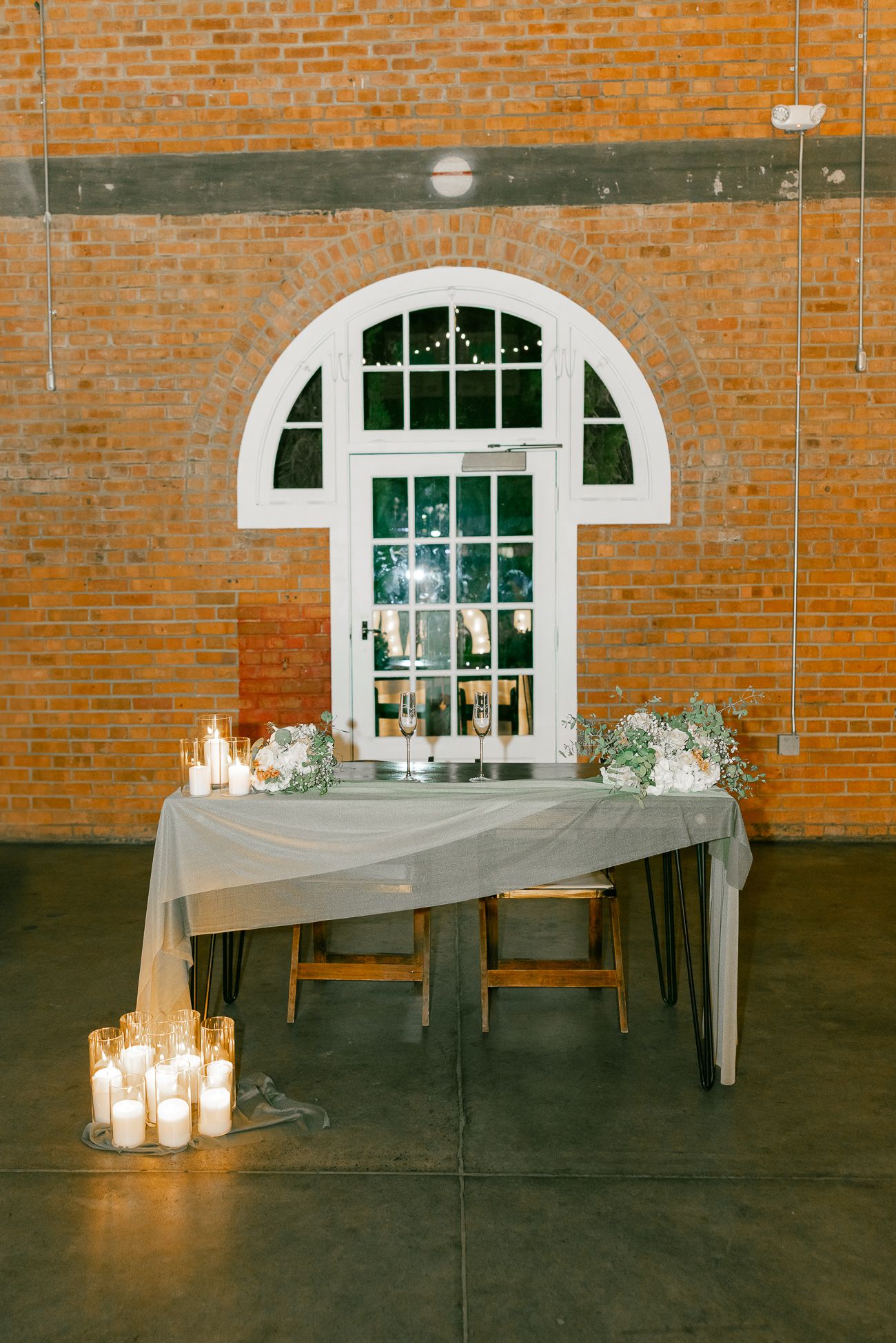 Wedding reception table setup with candles and floral arrangements against a brick wall with arched window.