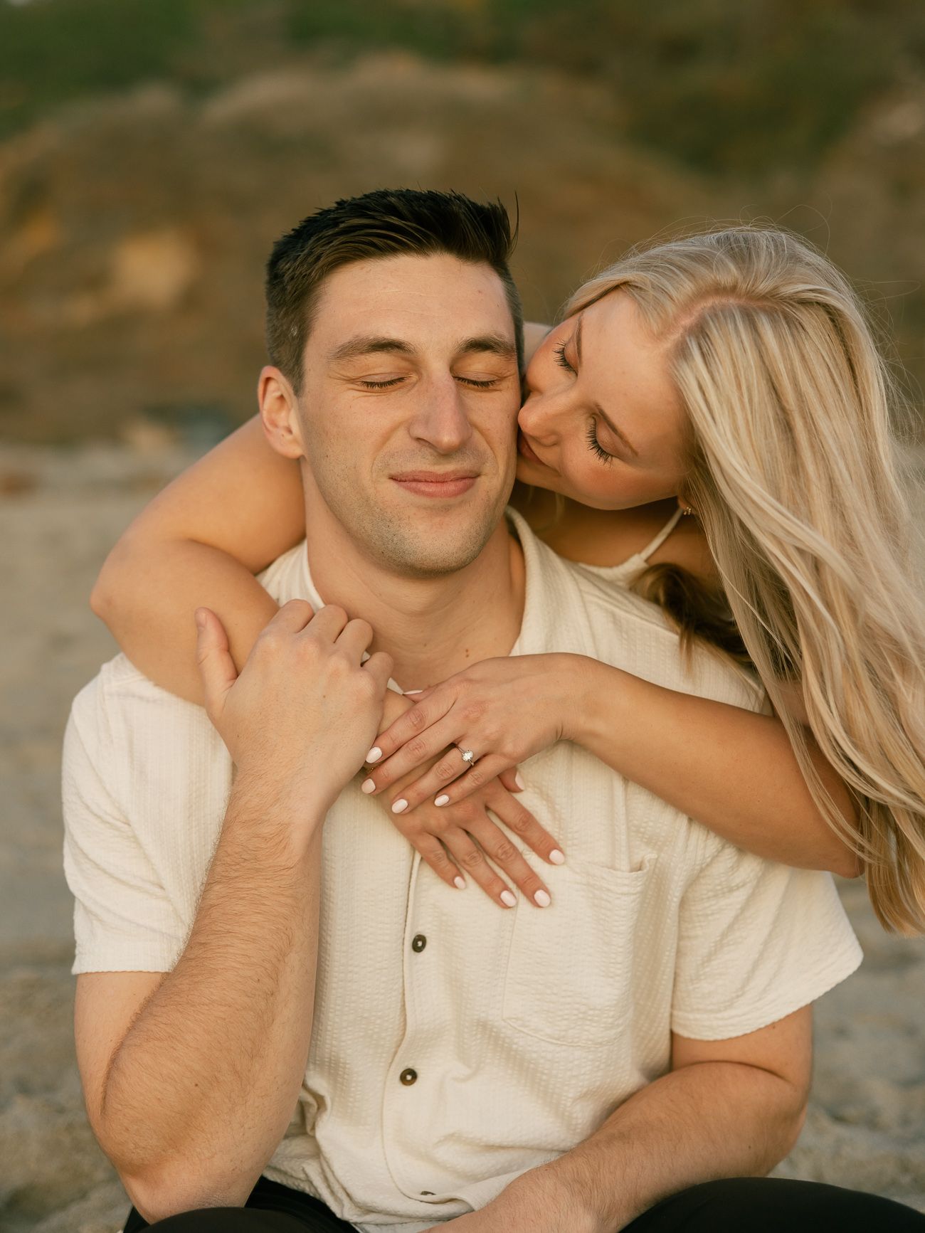 A romantic couple embraces outdoors at sunset, with the woman hugging from behind and showing affection in warm lighting.