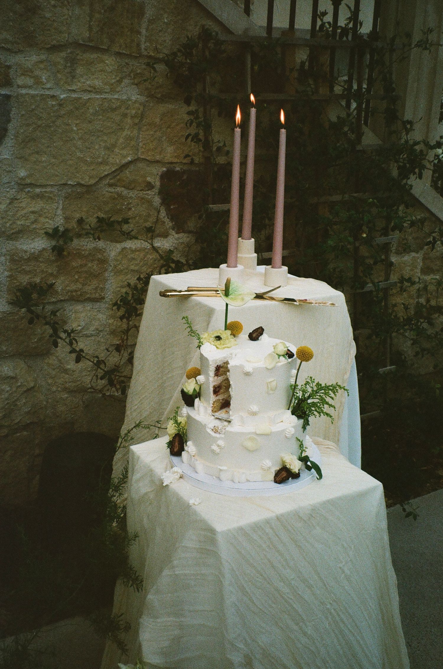 A white wedding cake adorned with tall candles and floral decorations displayed against a rustic stone wall.