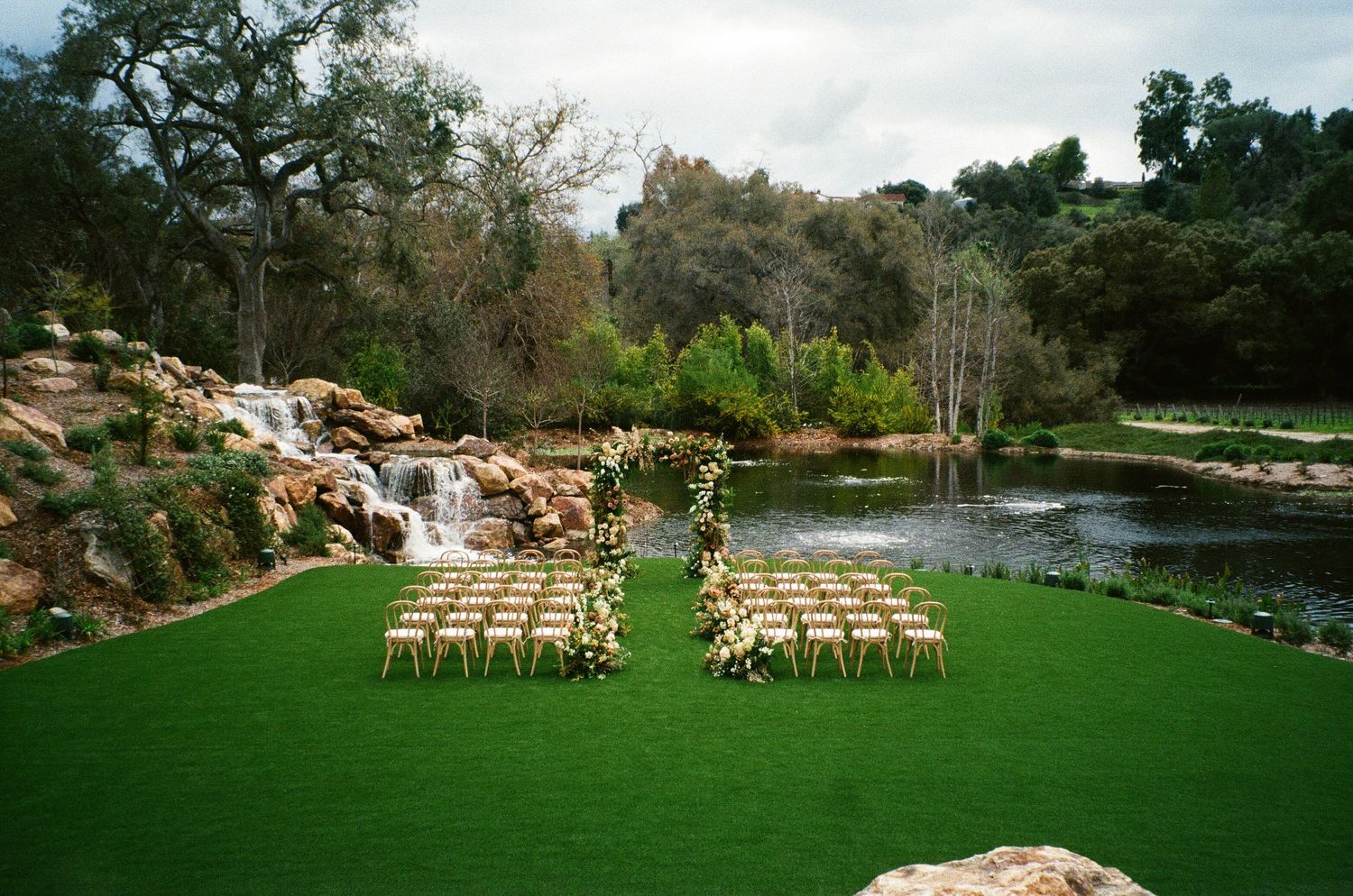 Outdoor wedding ceremony setup with wooden chairs on manicured lawn beside a waterfall and tranquil pond.