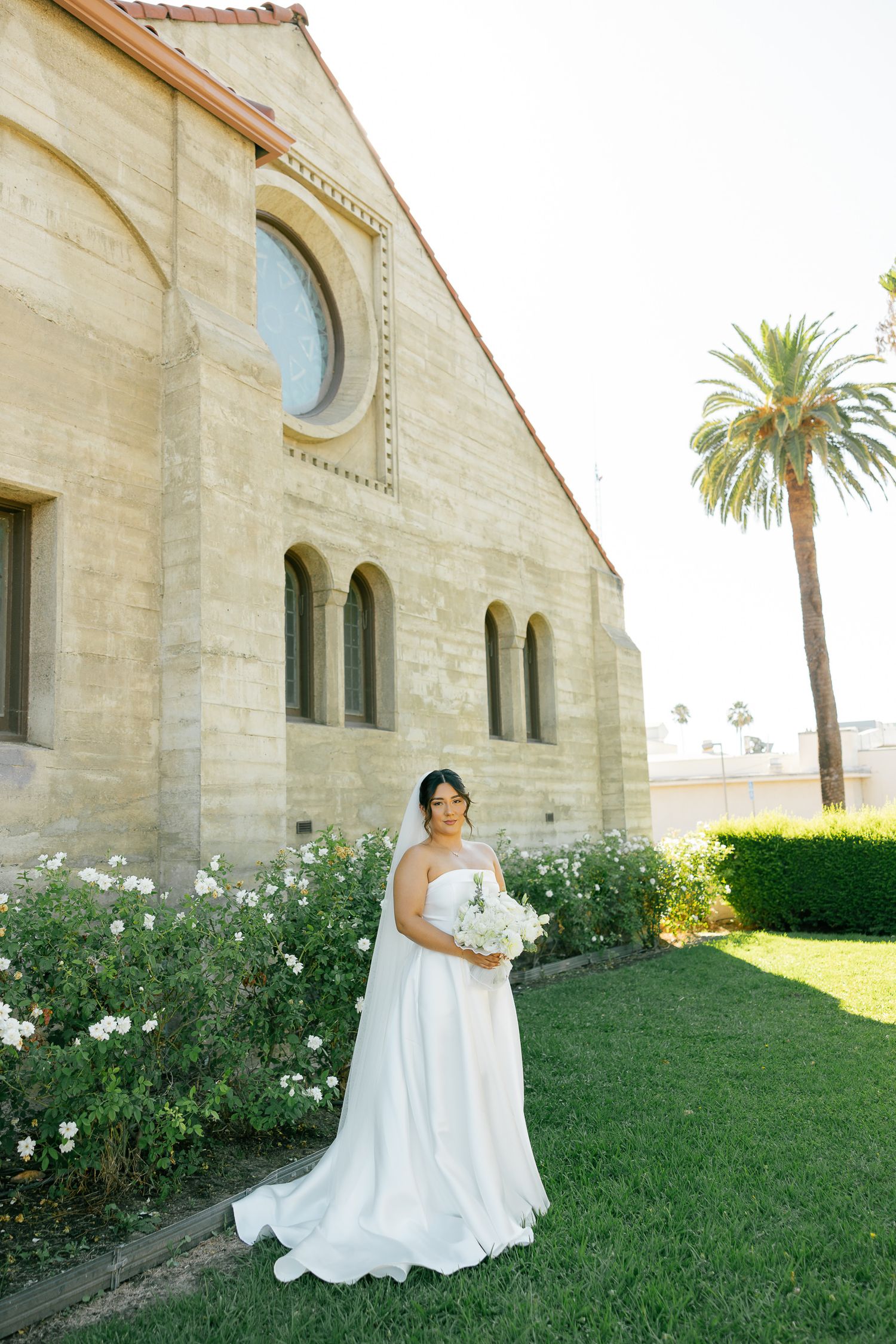 A white wedding gown stands out against a Spanish-style stone church with arched windows and a palm tree on a sunny day.