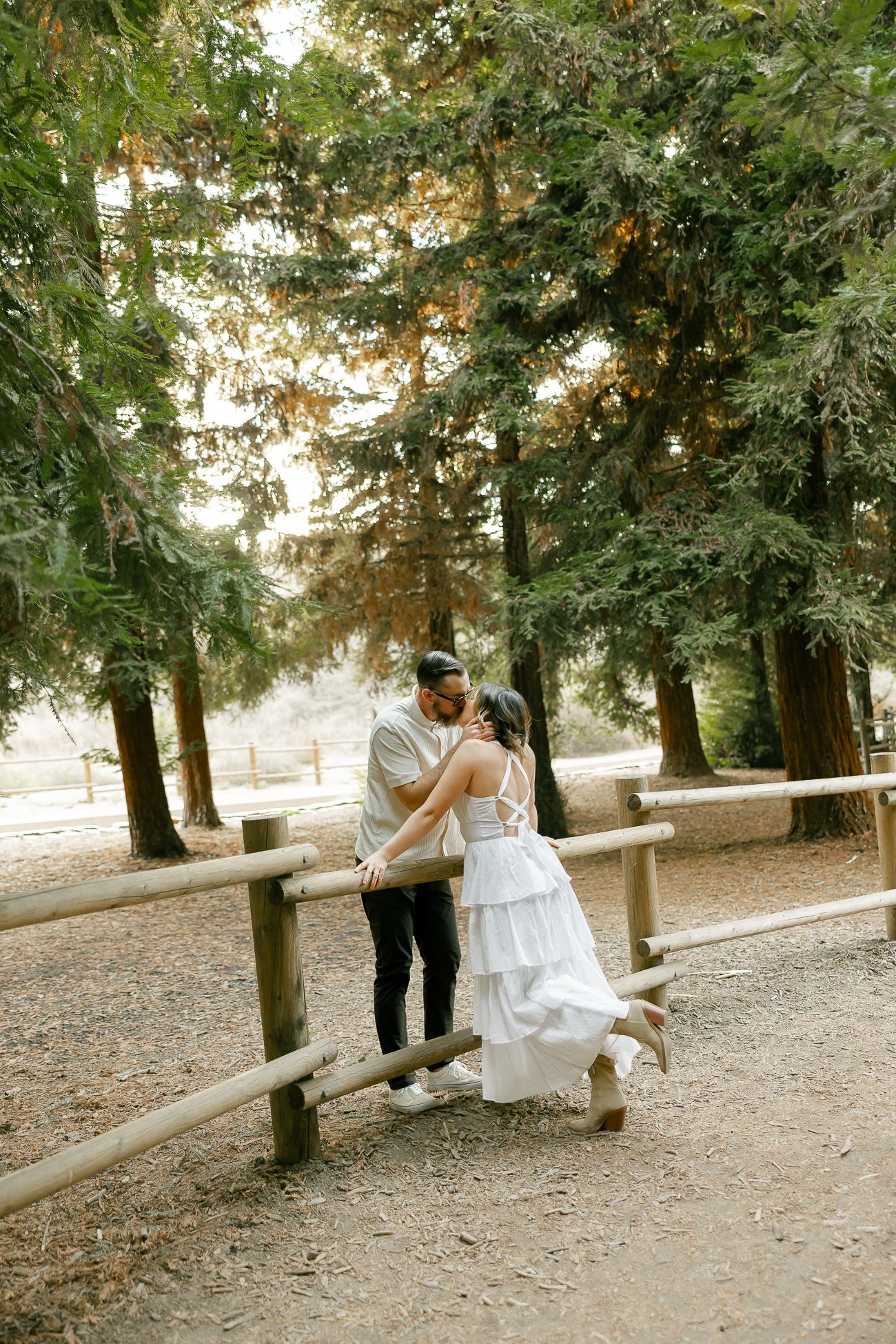 A couple shares a tender moment by a wooden railing in a serene forest setting.