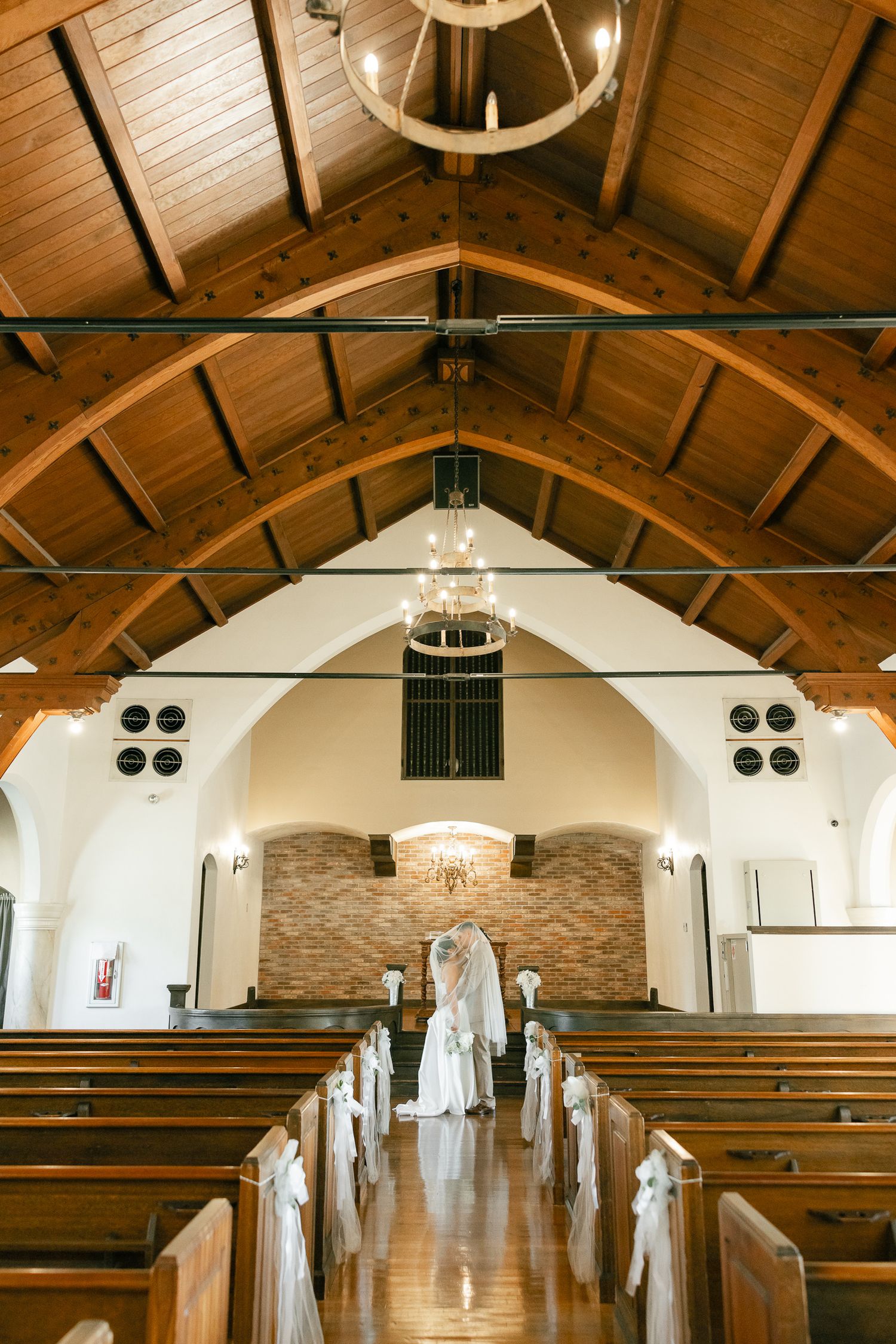 Interior view of a rustic church chapel with wooden beams, pews decorated with white bows, and stone accent wall at the altar.