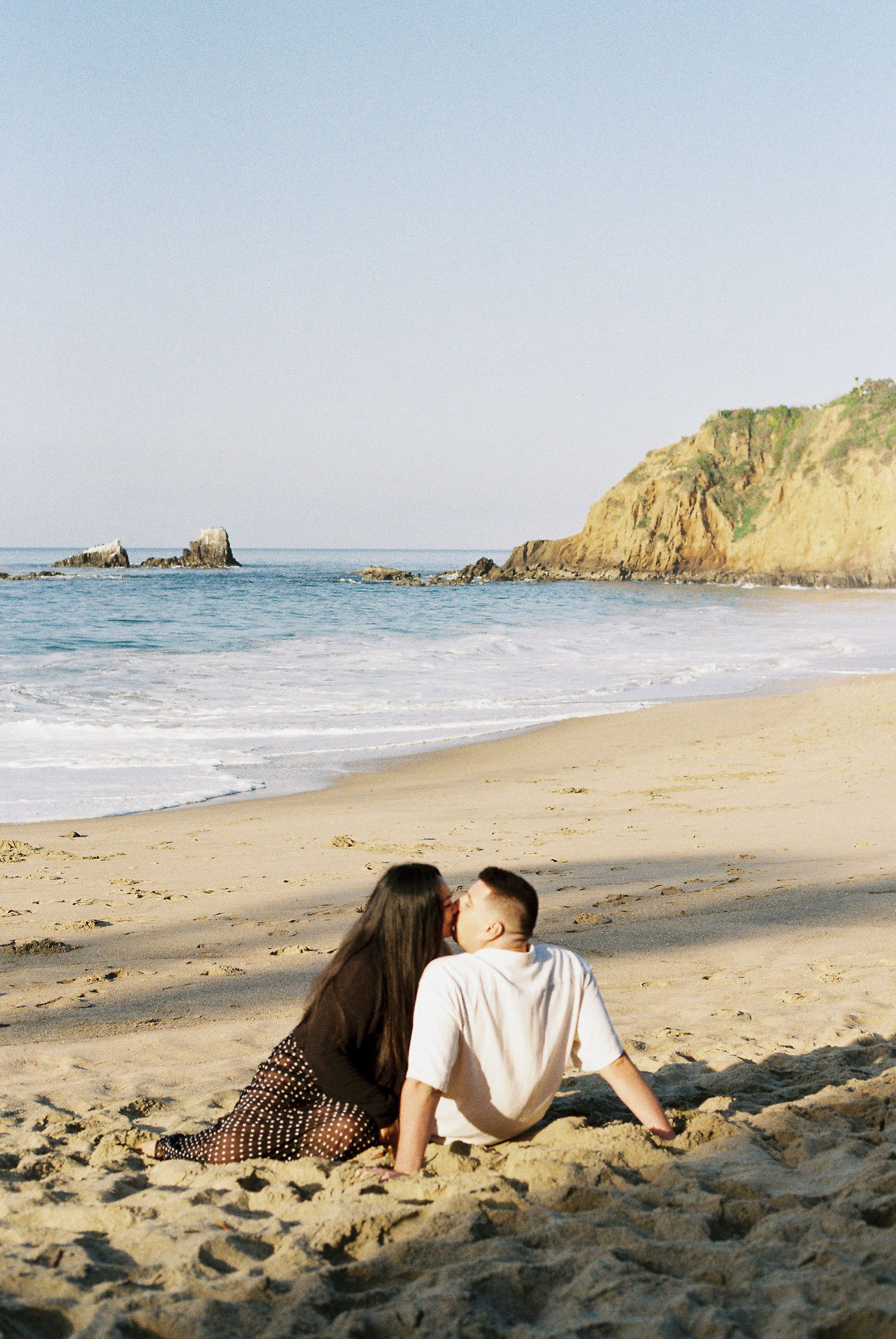 Two people relaxing together on a sandy beach with rocky coastal cliffs and ocean waves in the background.