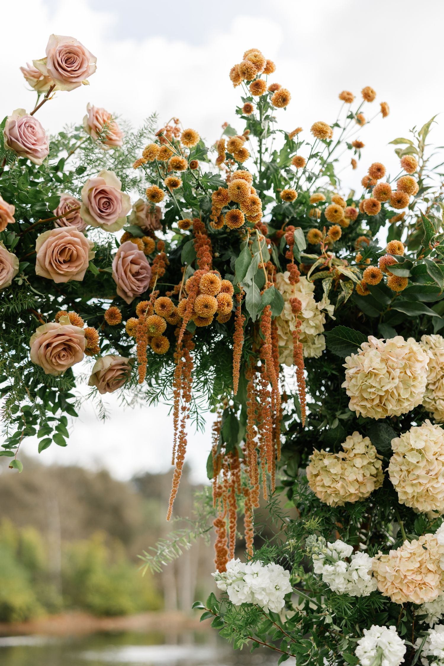 A lush floral arrangement with blush roses, orange blooms, and white hydrangeas cascading against a natural backdrop.