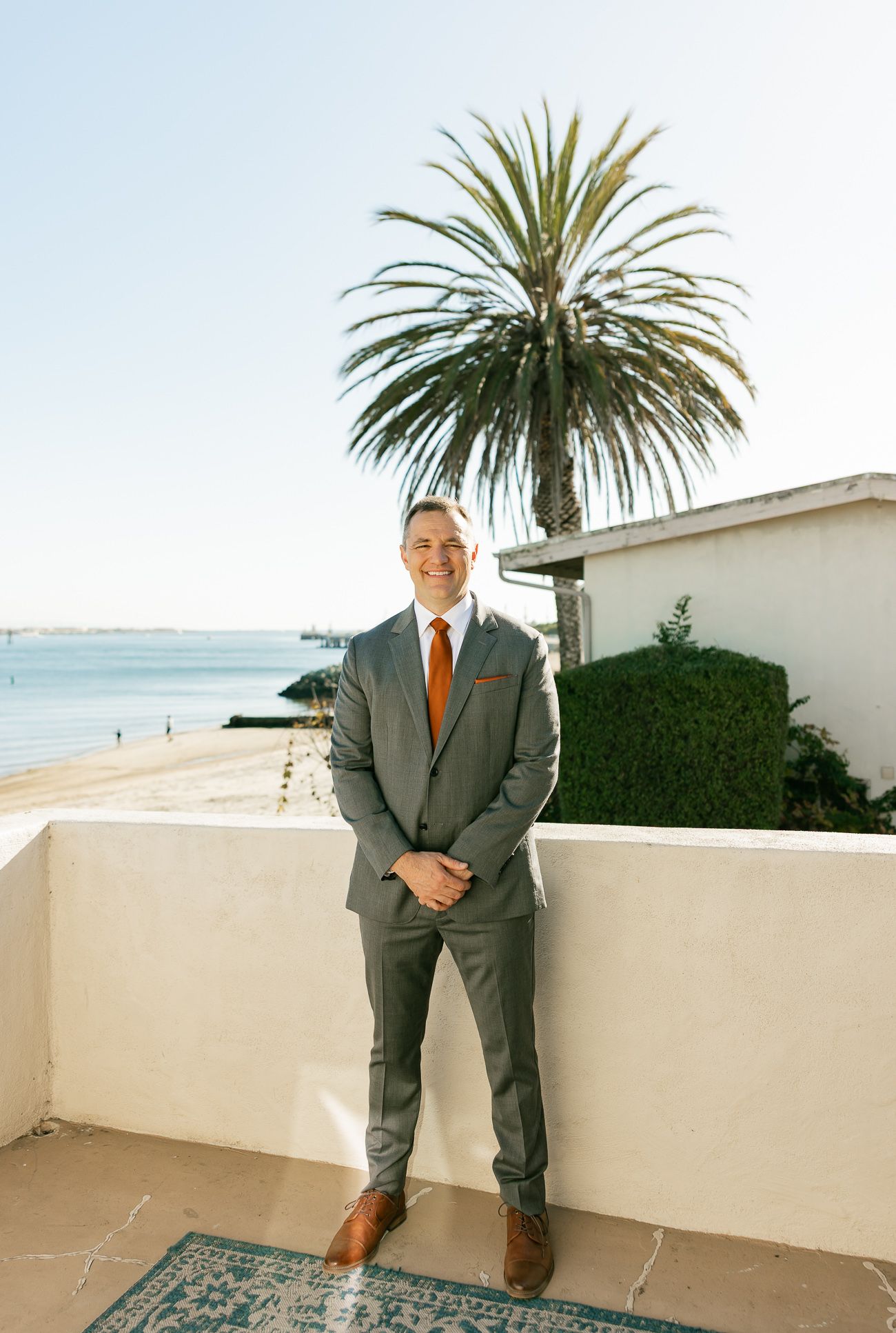 Groom in grey suit and orange tie poses by palm tree on coastal terrace.
