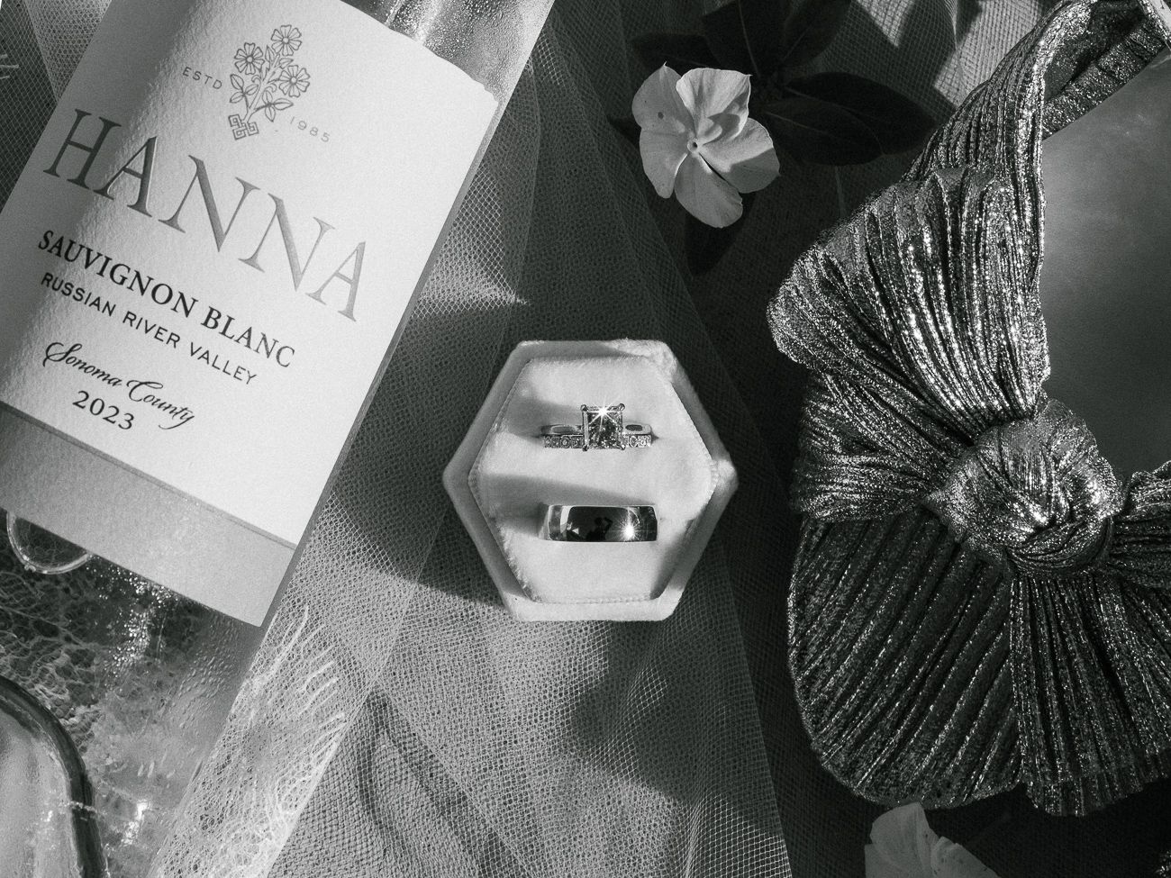 A monochromatic still life featuring a wine bottle, hexagonal ring box, and textural decorative elements in black and white.