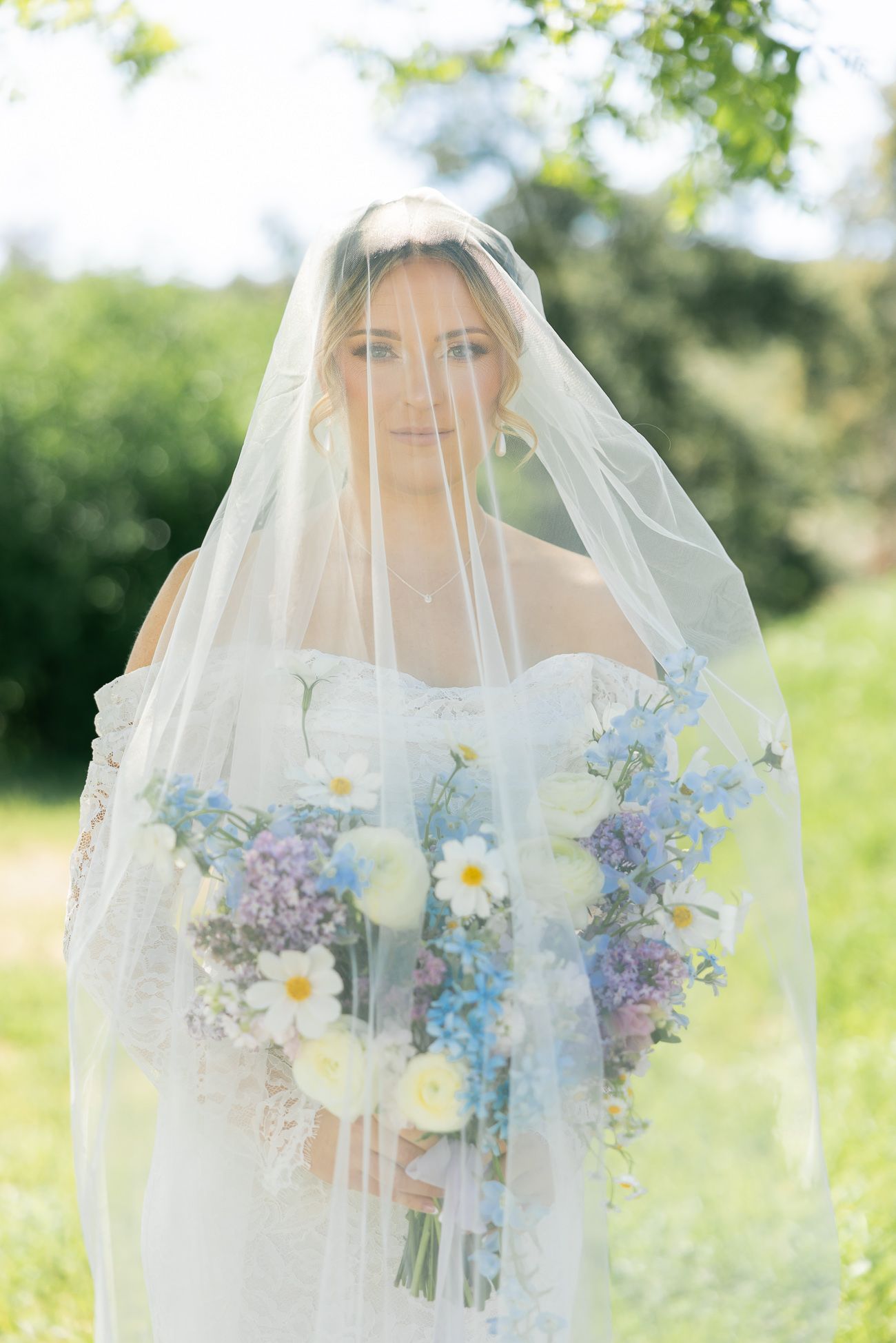 Bride in white veil holds stunning bouquet of blue delphinium, daisies and purple flowers in sunny garden setting.