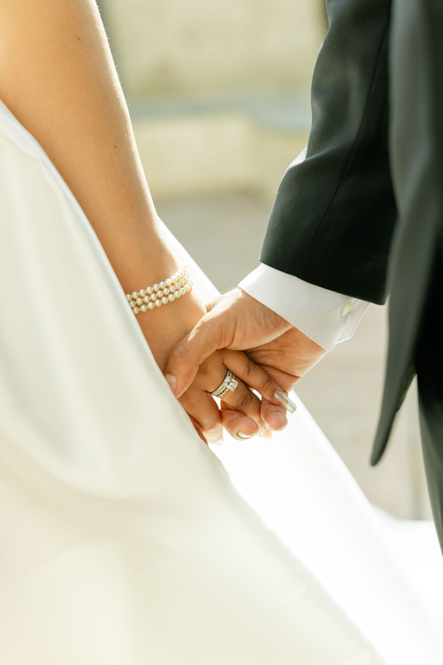 Close-up of bride and groom holding hands, showing wedding rings and pearl bracelet against white dress and black suit.