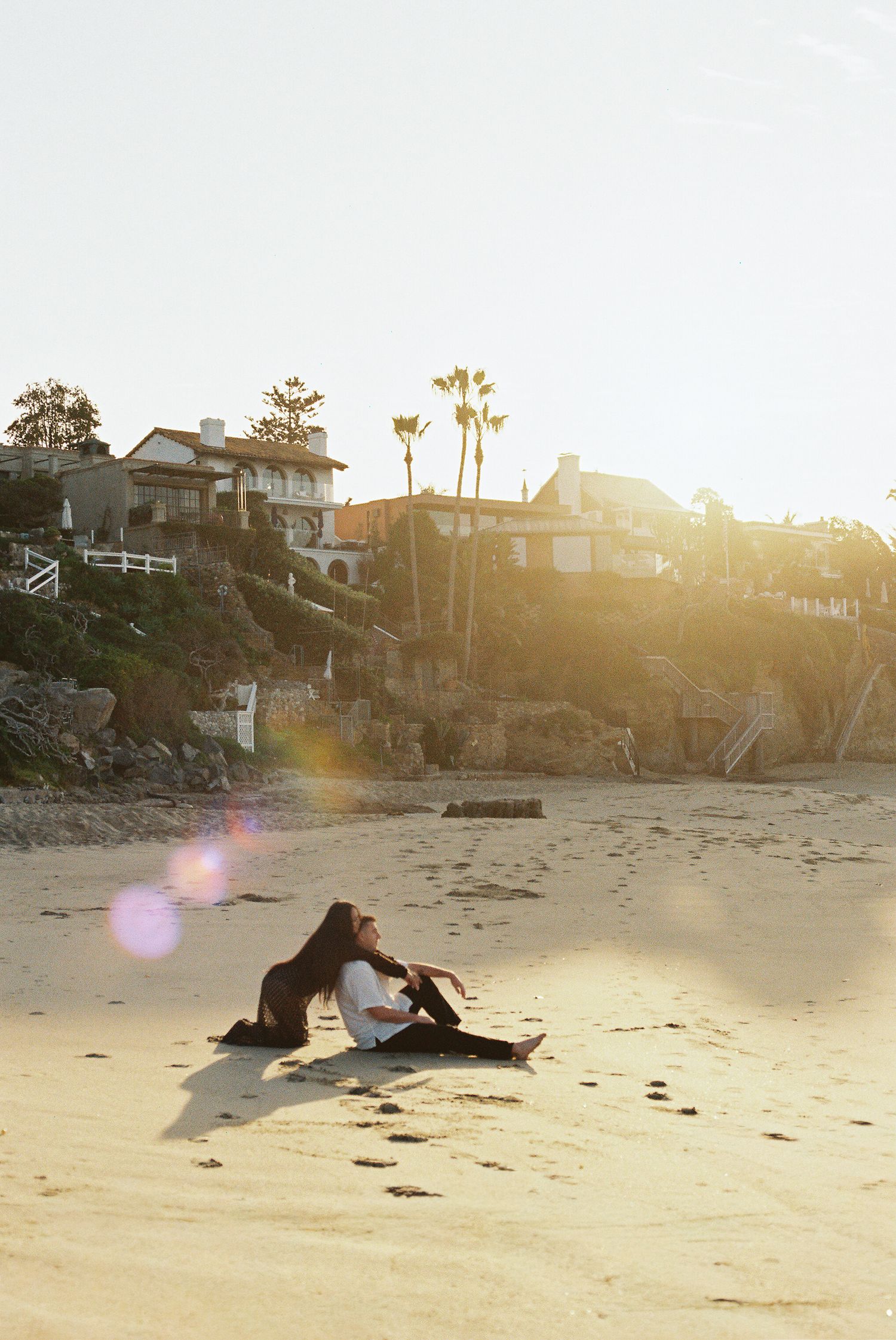 People relaxing on a sandy beach at sunset with palm trees and beachfront homes in the background.