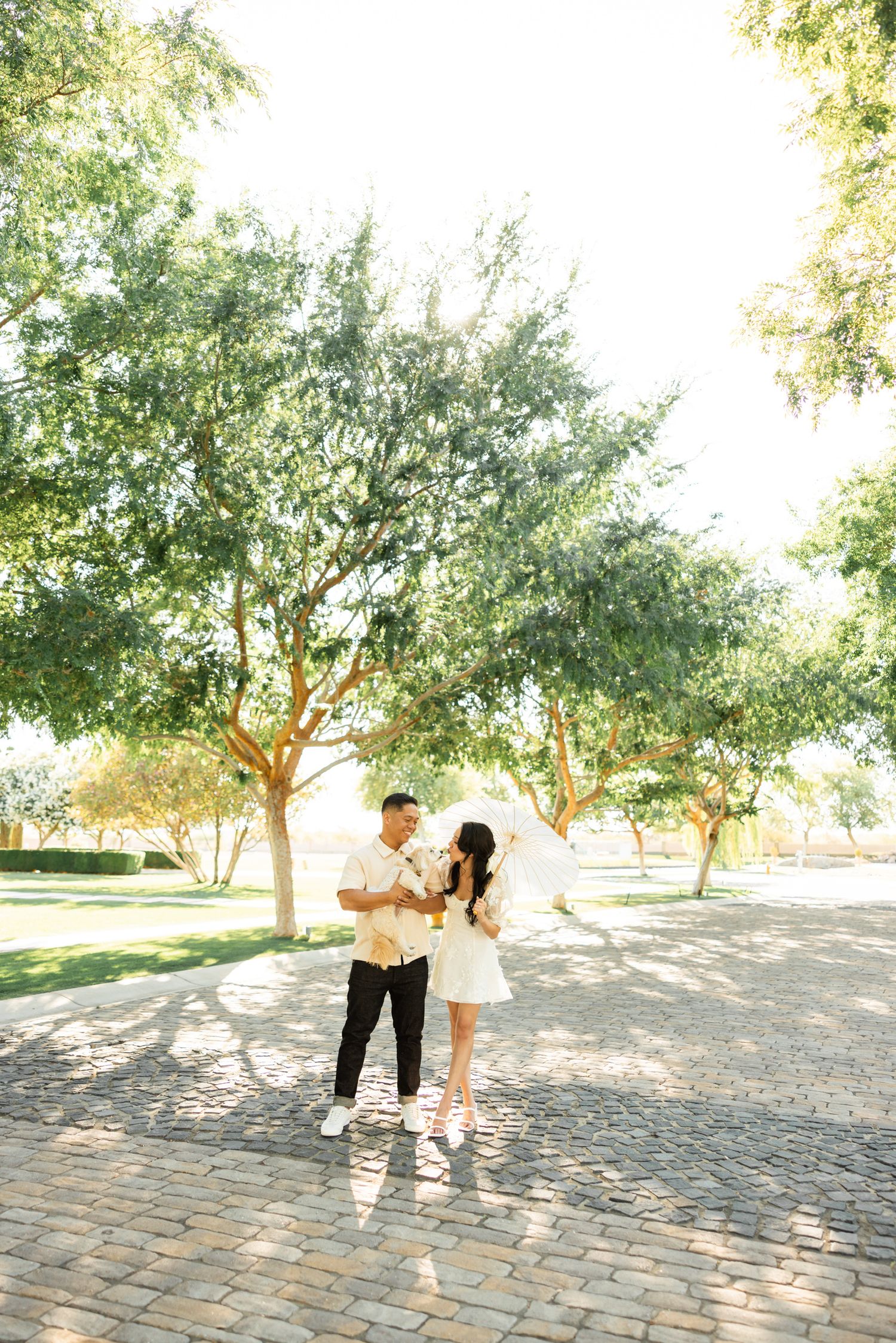 A couple dances playfully on a brick patio surrounded by trees in a romantic outdoor setting.