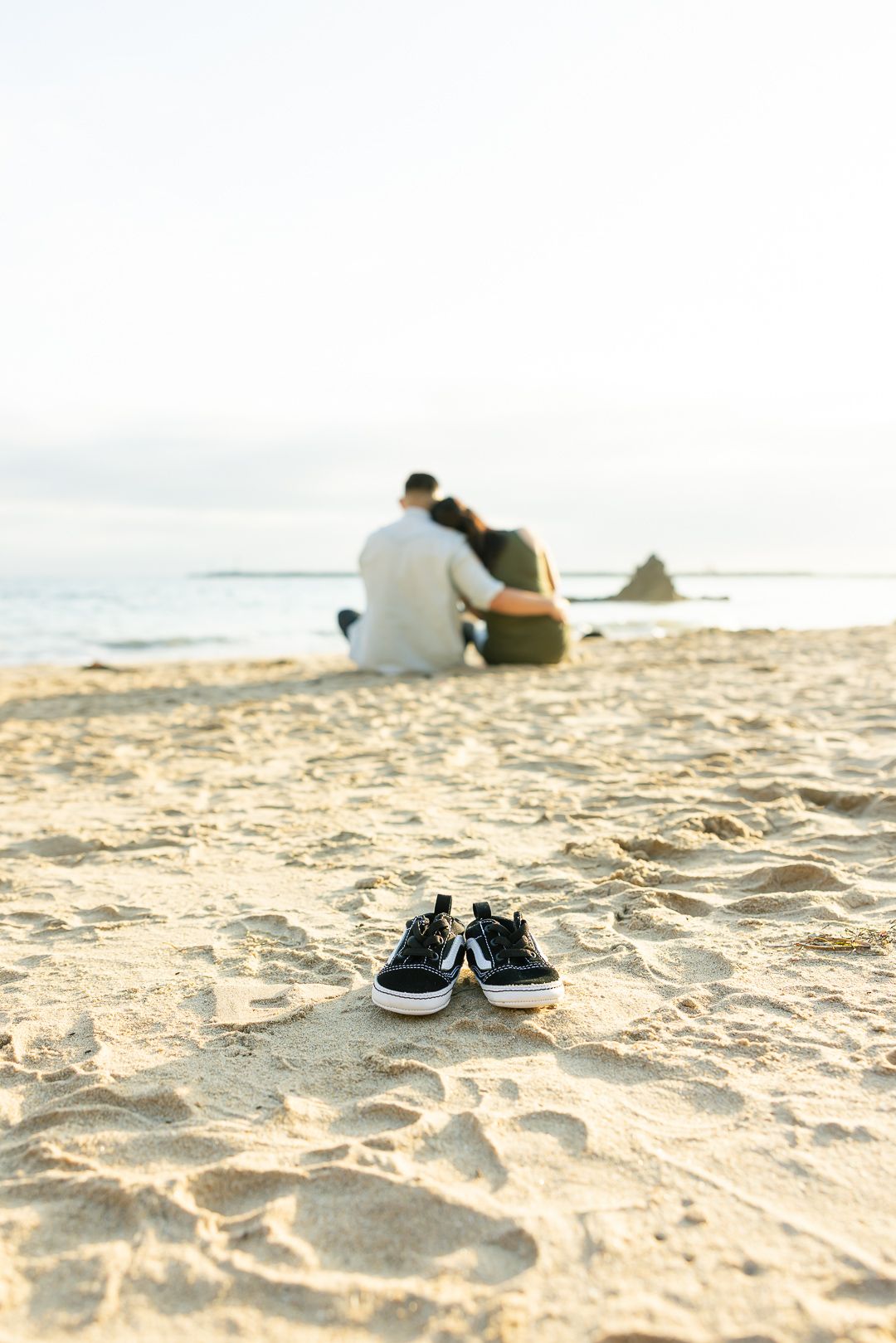 A pair of navy blue sneakers in the sand with a couple sitting by the ocean in soft sunlight.