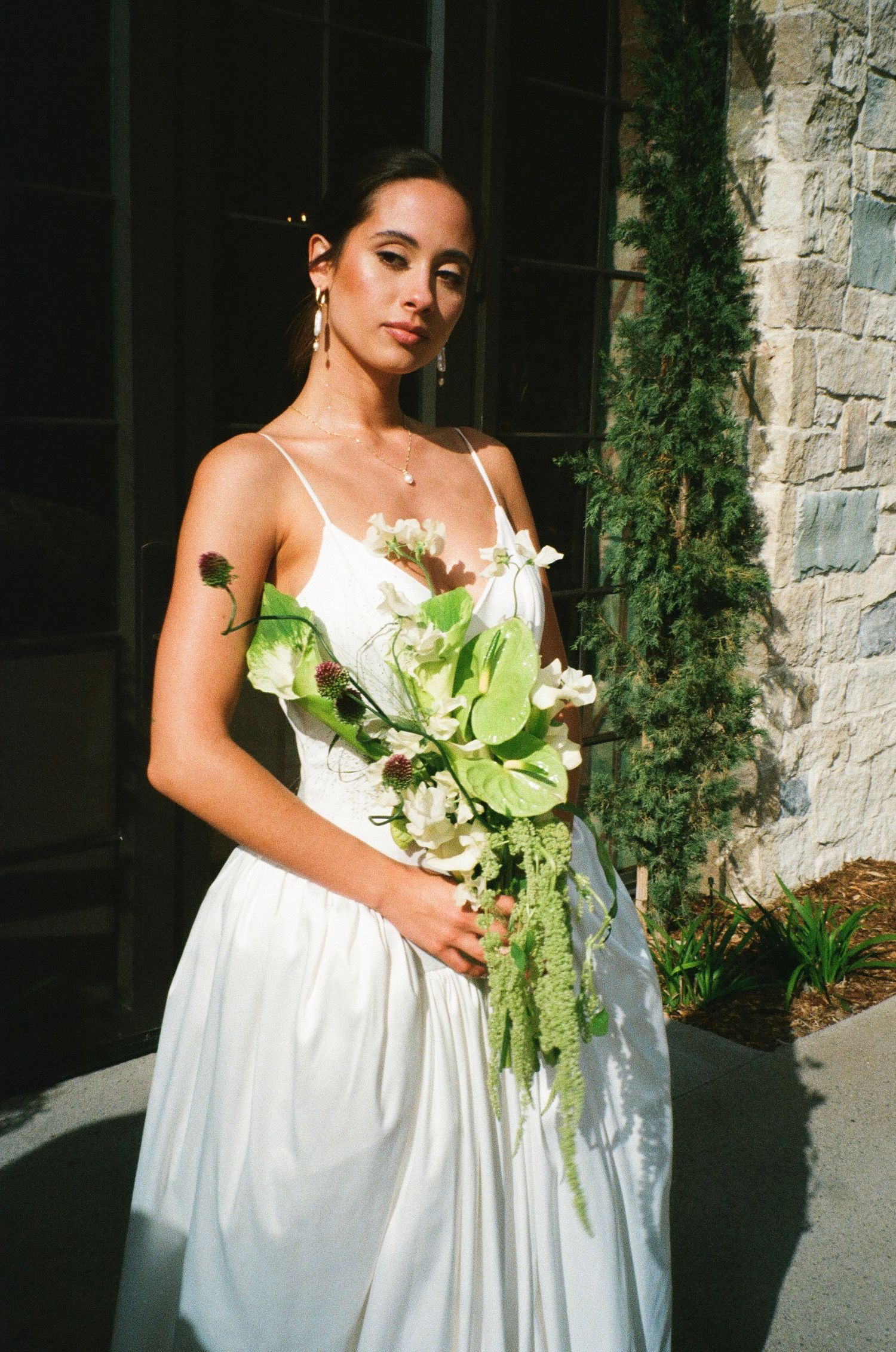 A bride in a white satin gown holds a unique green and white floral bouquet against a stone wall backdrop.