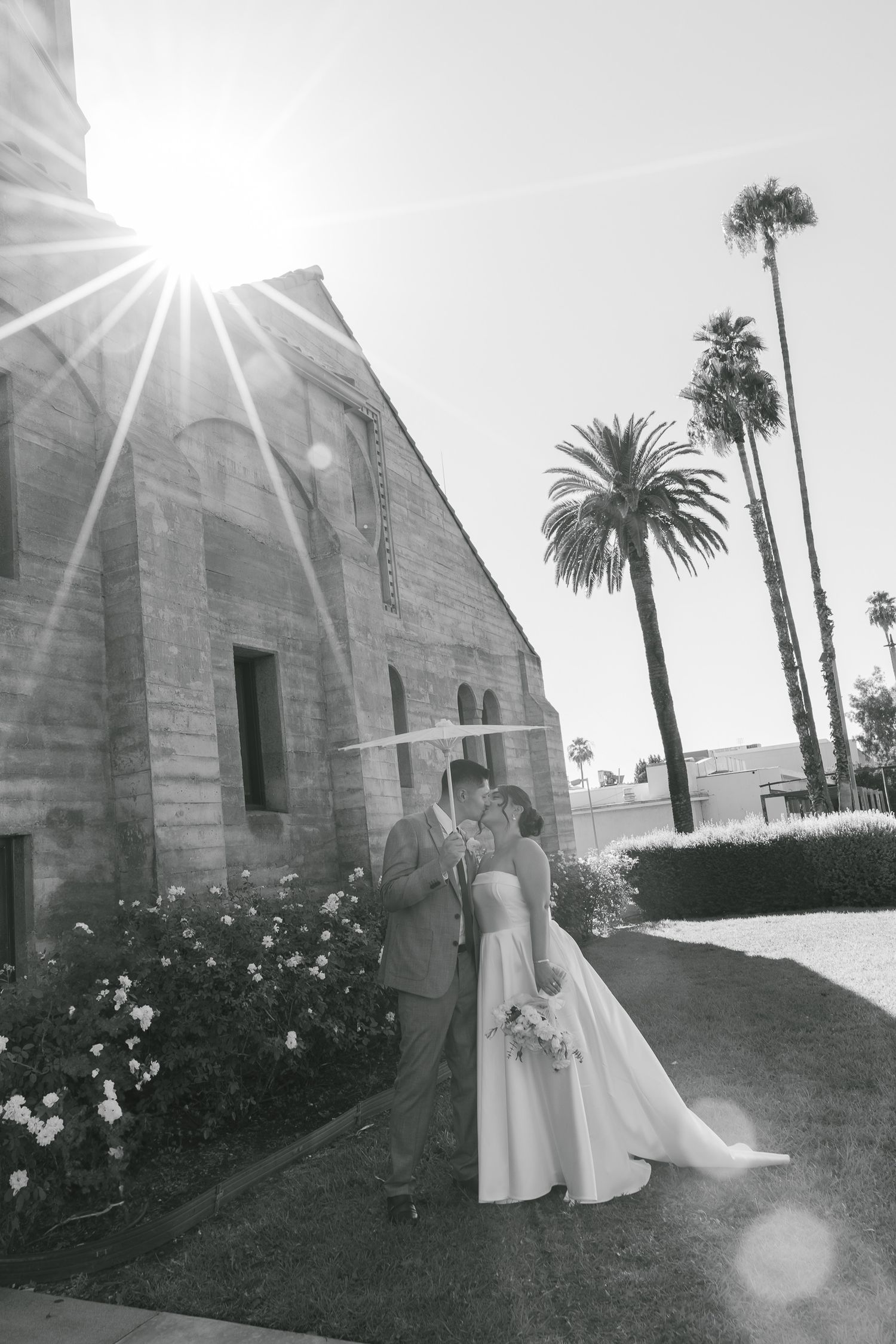 A romantic wedding scene outside a stone church with tall palm trees and bright sunlight.
