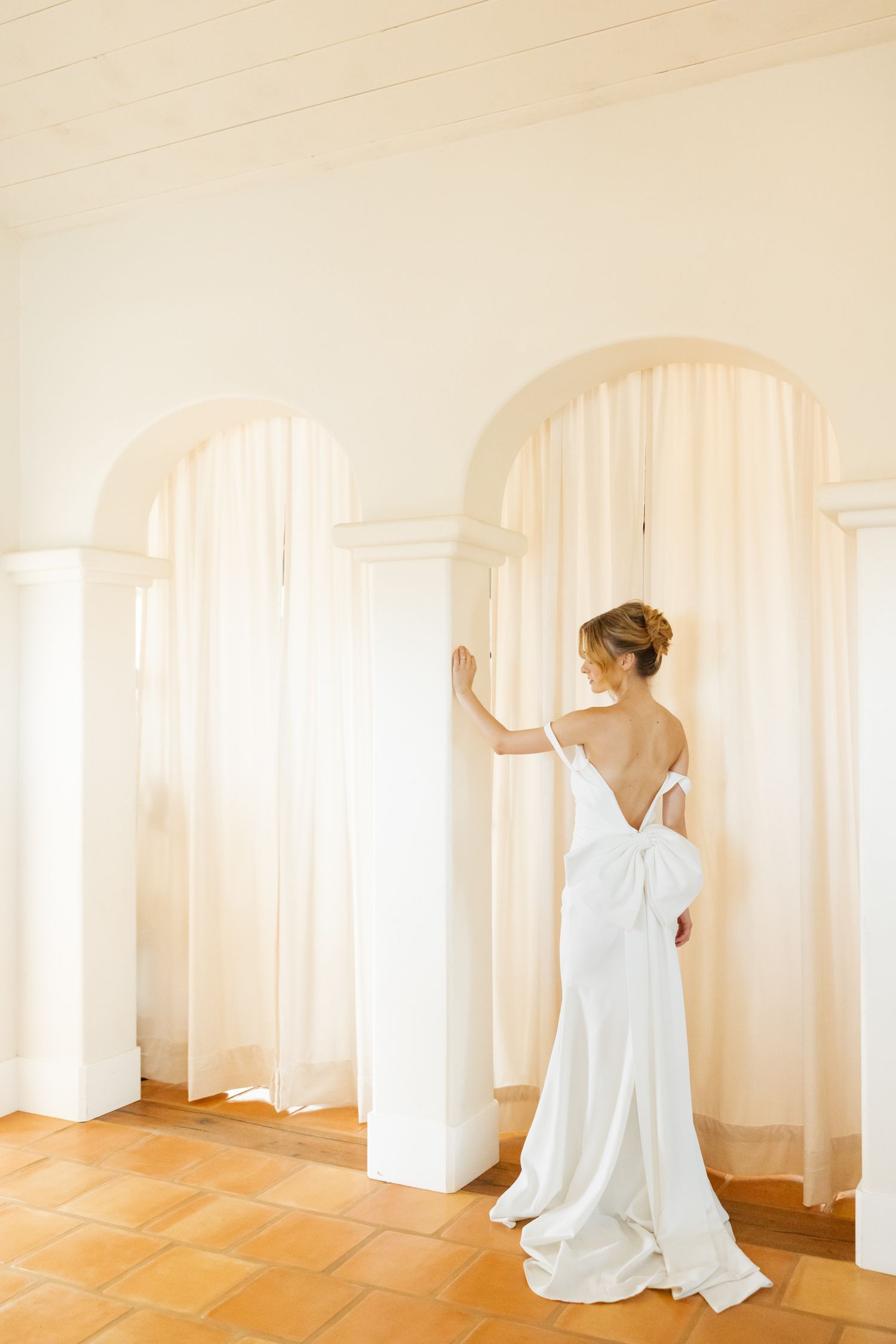 Elegant bride in flowing white backless gown poses near Mediterranean-style archways and terra cotta floor.