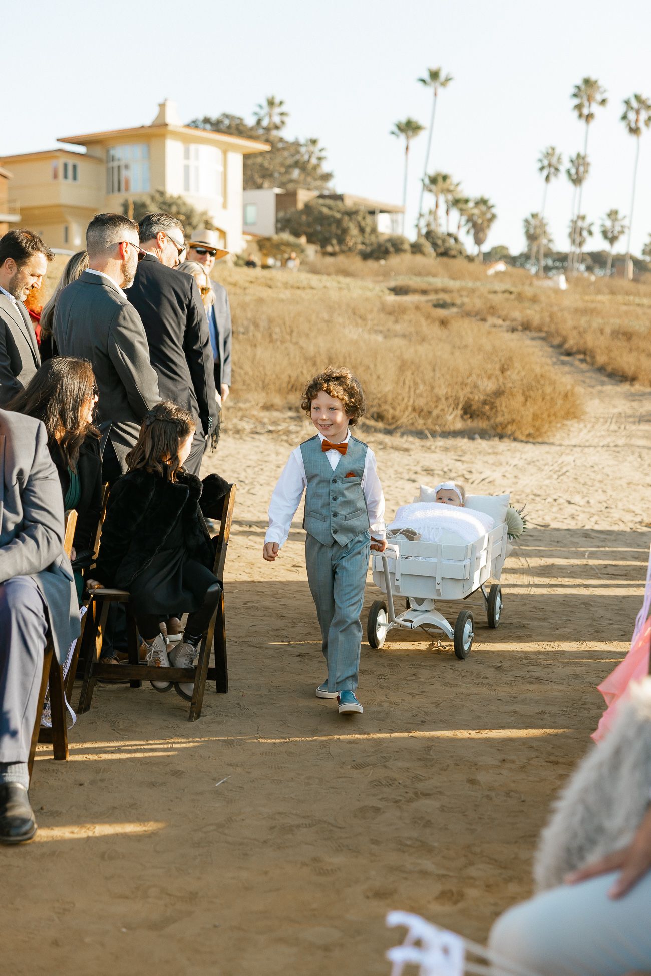A child in a blue vest walks on a beach path while carrying a small wagon during a gathering of formally dressed guests.