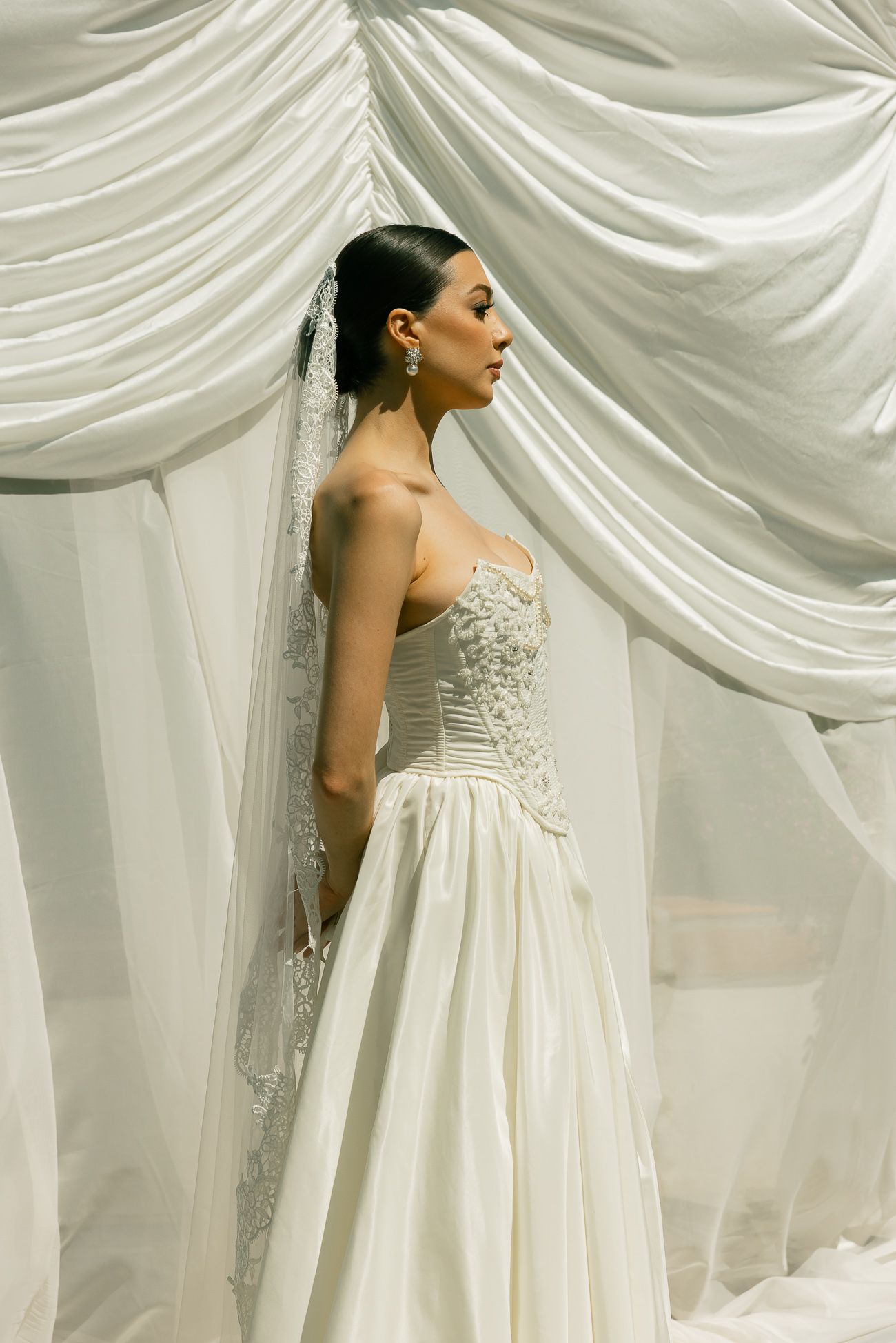 A bride in an elegant white strapless wedding gown with lace details stands against draped white fabric backdrop.