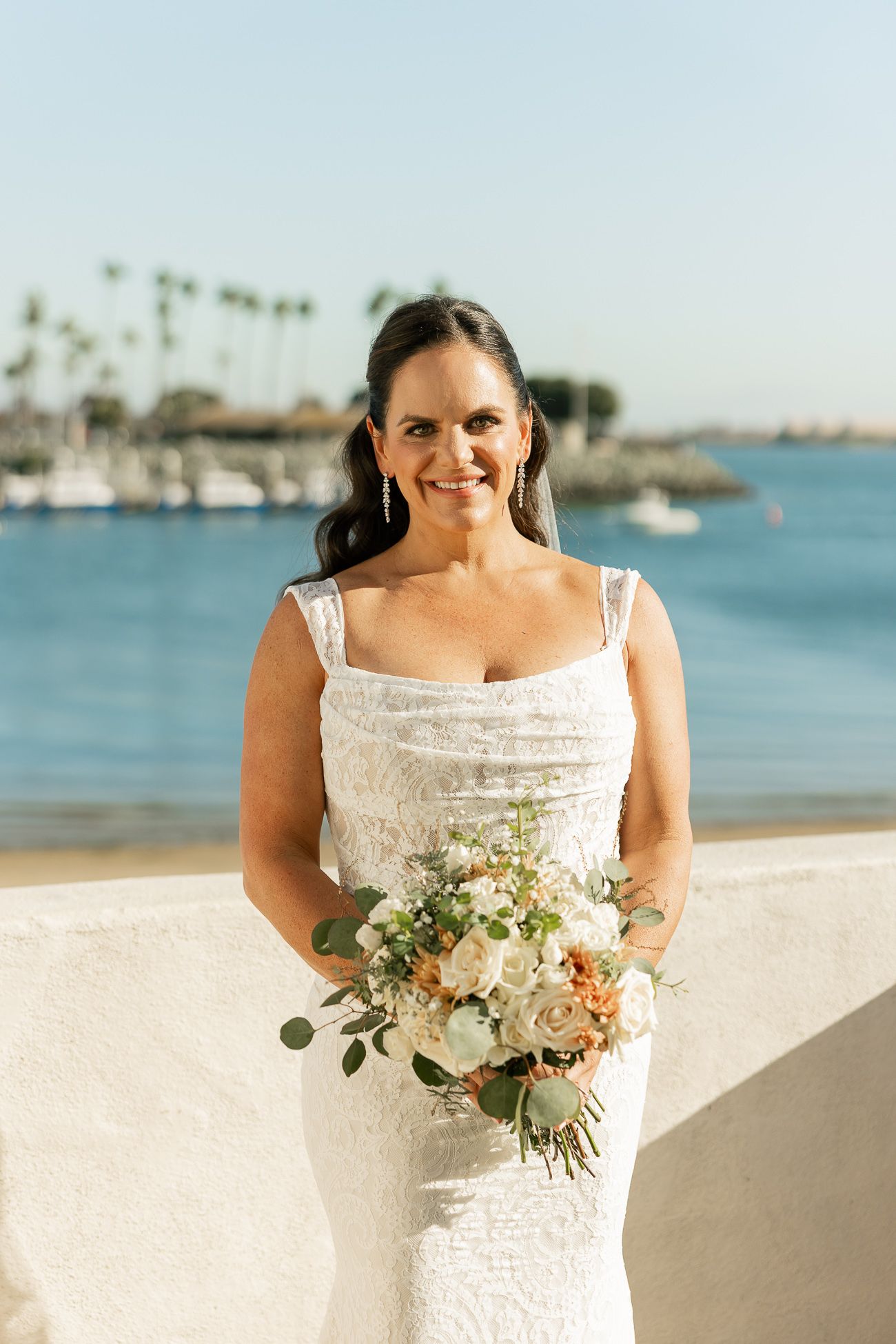 Beautiful beachfront wedding with palm trees and marina in background, featuring white lace dress and soft floral bouquet.