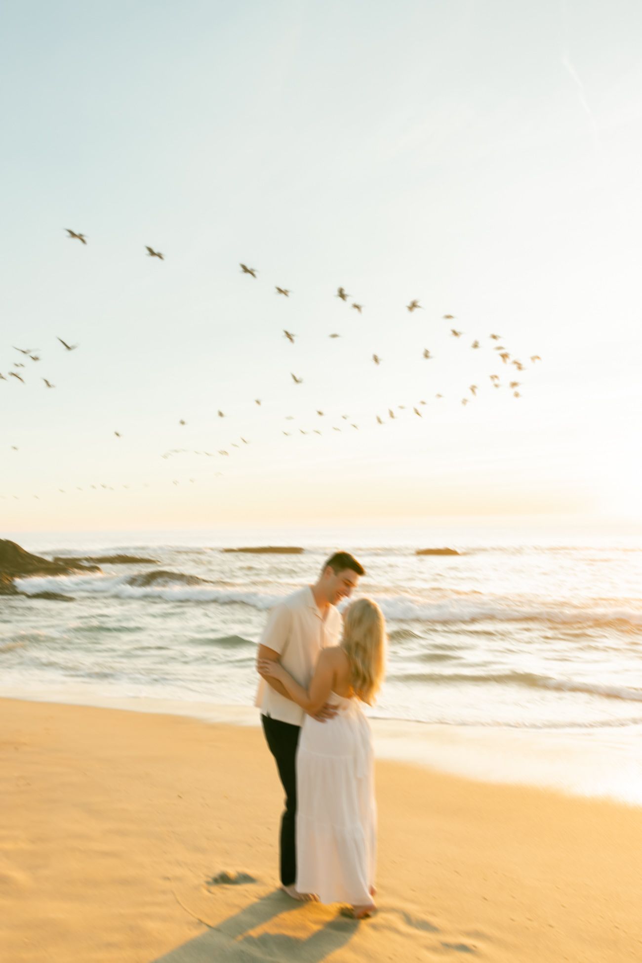 A couple embraces on a sunset beach while birds soar overhead in a dreamy golden light.