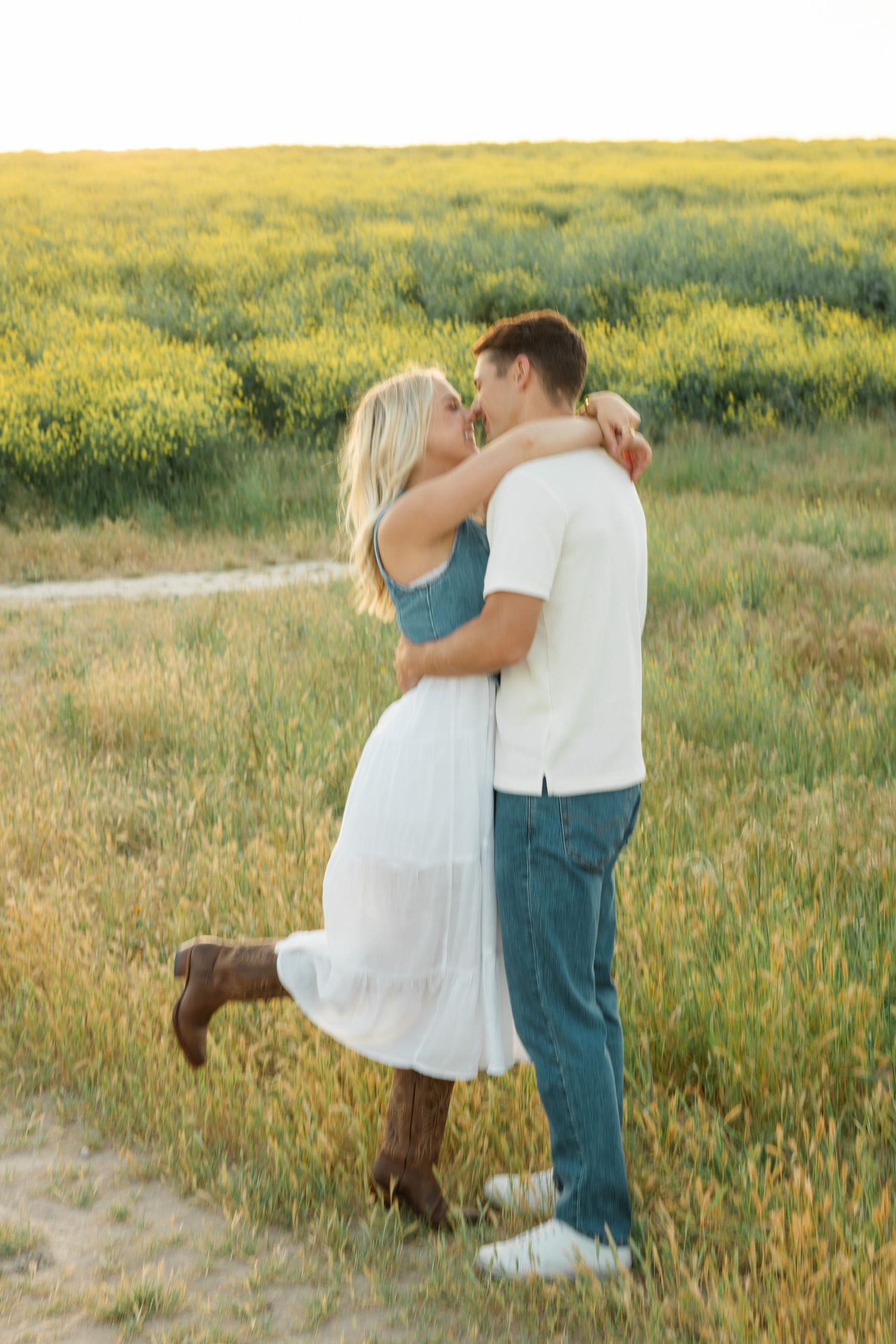 A couple embraces while dancing on a dirt path surrounded by golden wildflowers at sunset.