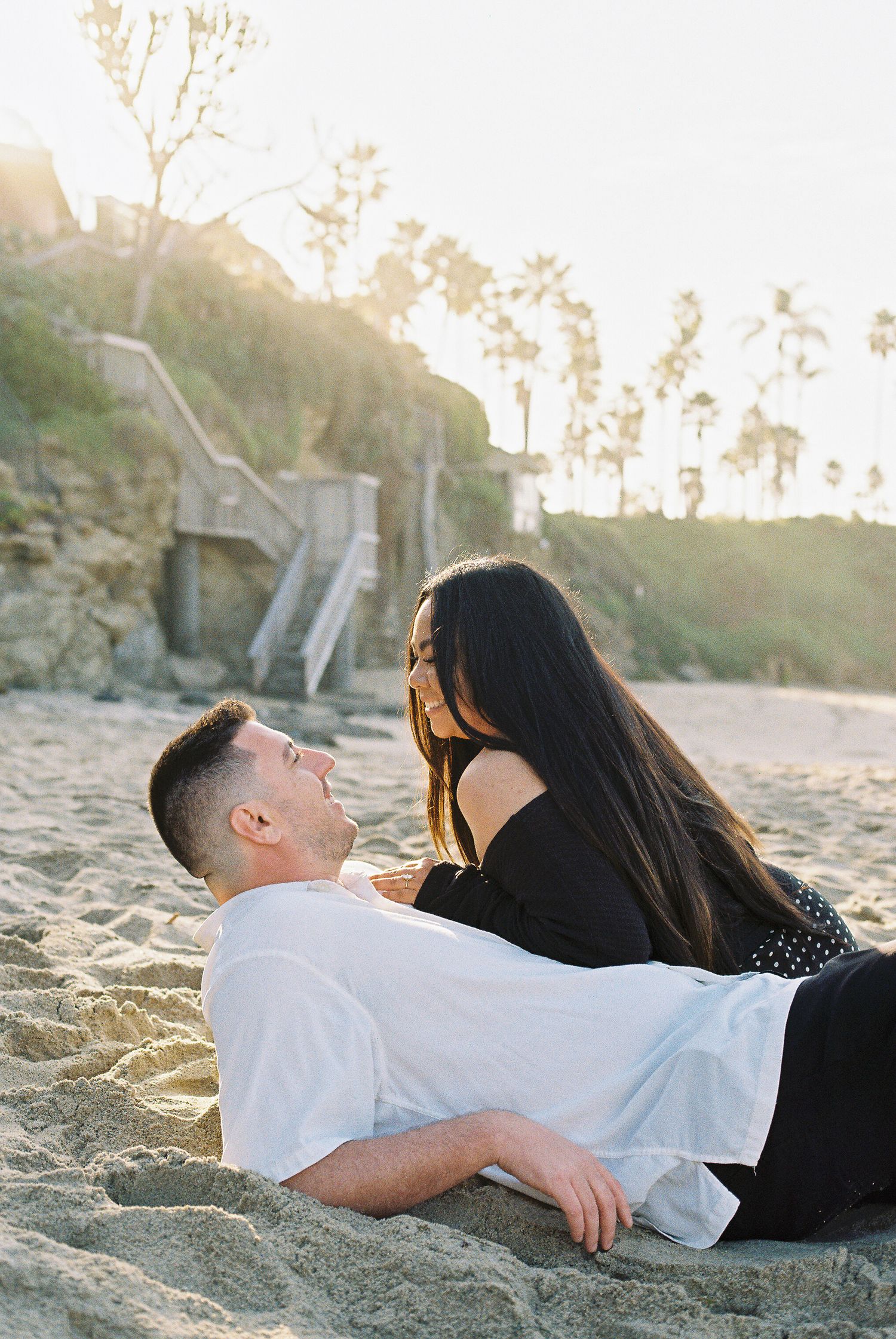 A romantic couple lying together on a beach at sunset with palm trees in the background.
