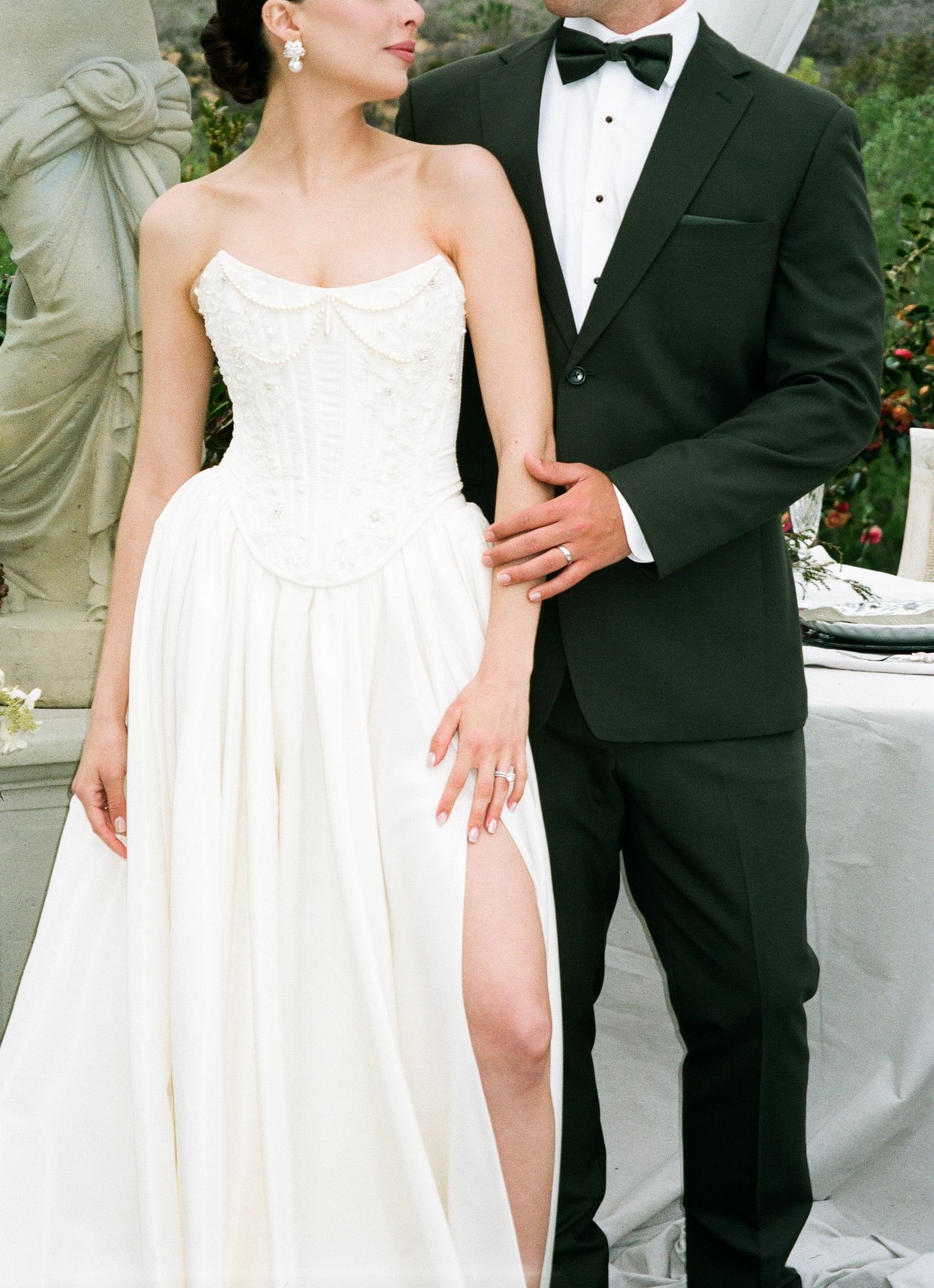 A bride in a flowing white gown and groom in black tuxedo pose together in an elegant garden setting.