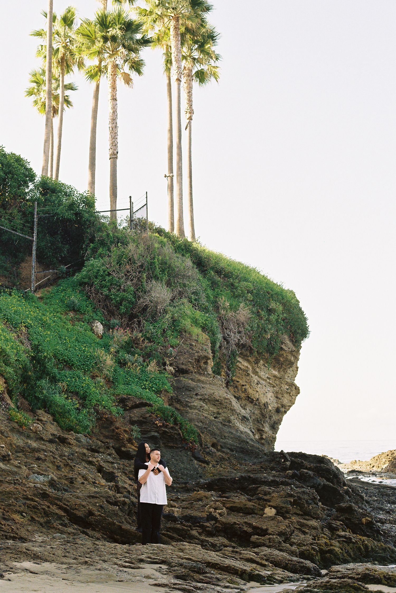 Rocky coastal cliff with tall palm trees silhouetted against bright sky at sunset.