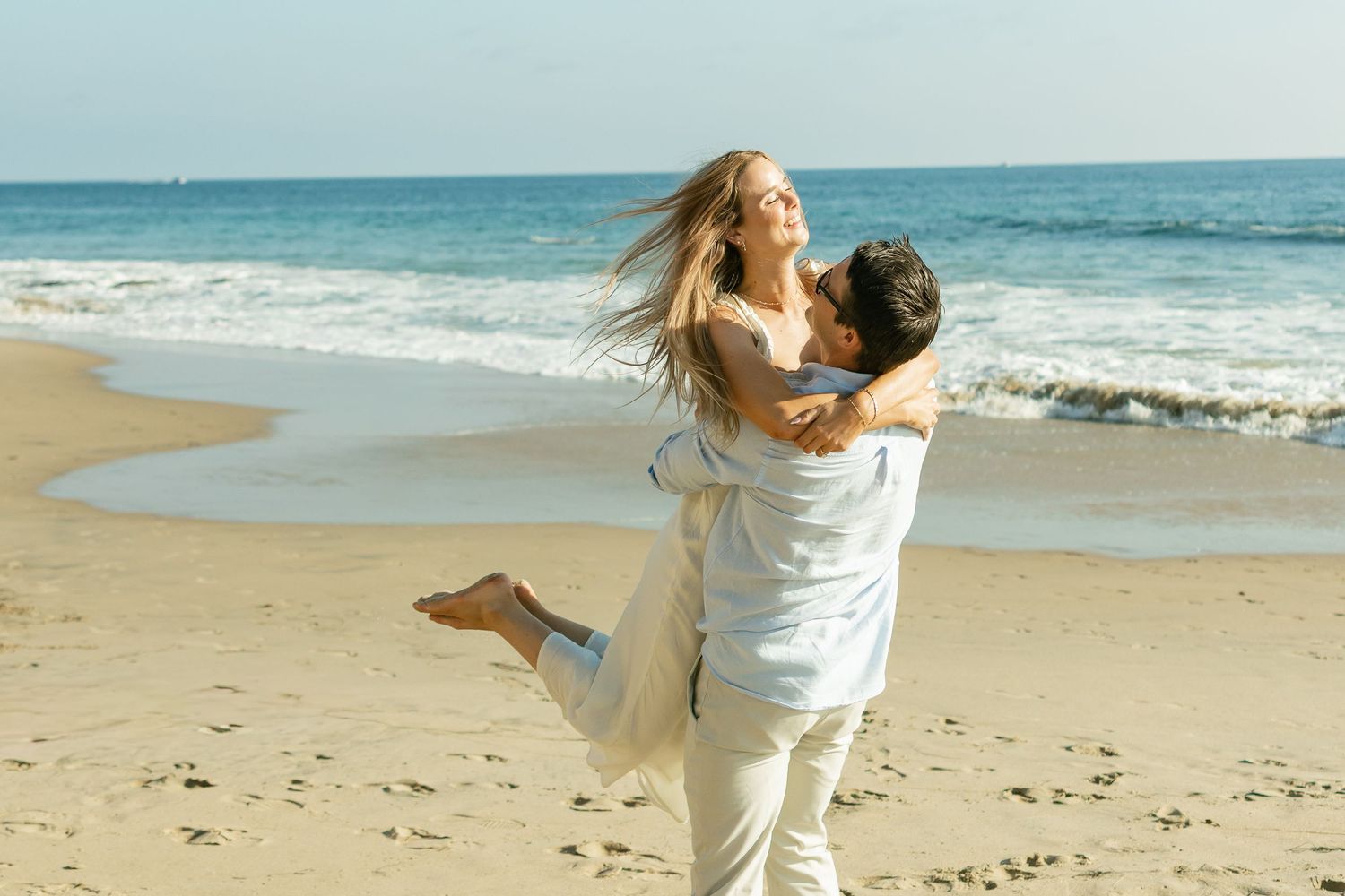 A person in white clothing spins joyfully on a sunny beach with ocean waves in the background.