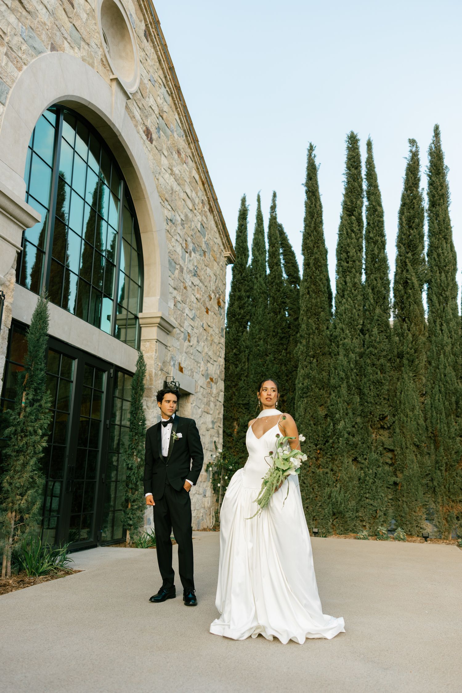 A couple in wedding attire stands outside a stone building with arched windows and tall cypress trees in the background.