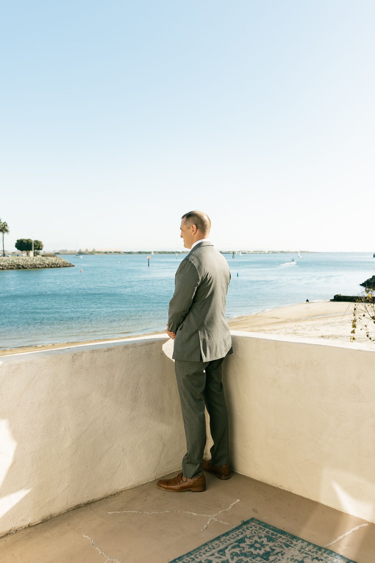 Person in gray suit looks out over a scenic beachfront balcony with blue ocean waters and palm trees in the distance.