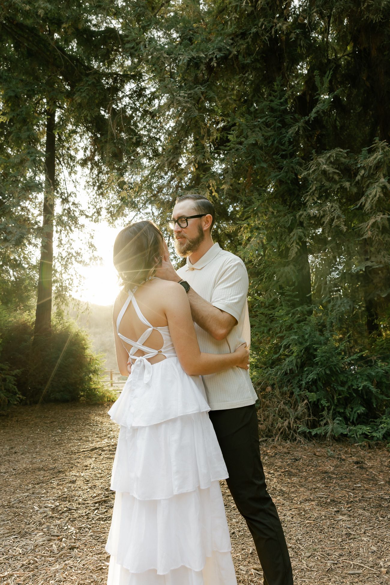 A romantic couple embraces in a sunlit forest setting with the bride wearing a flowing white tiered dress.