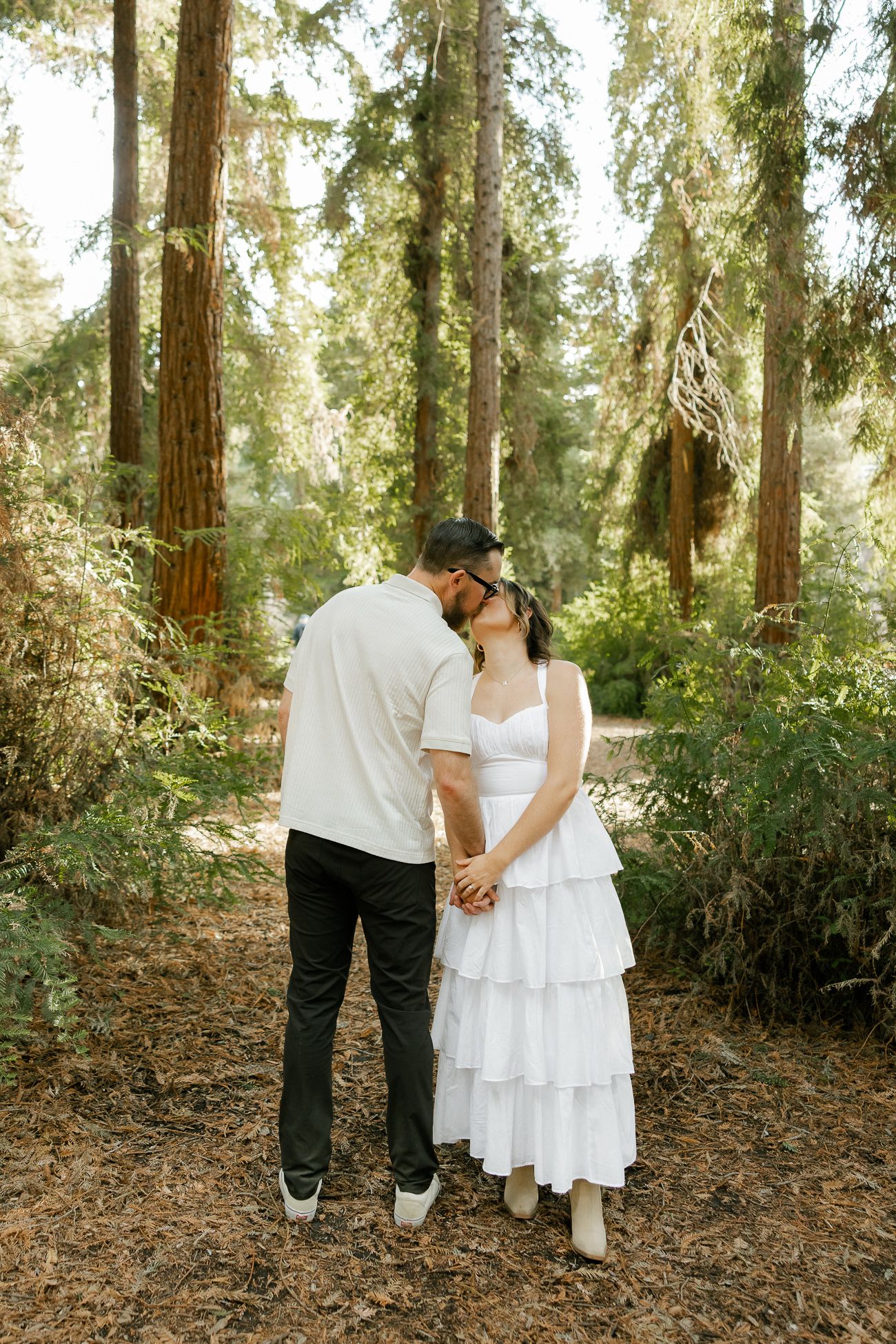 A couple shares an intimate moment while holding hands in a sunlit forest with tall redwood trees in the background.