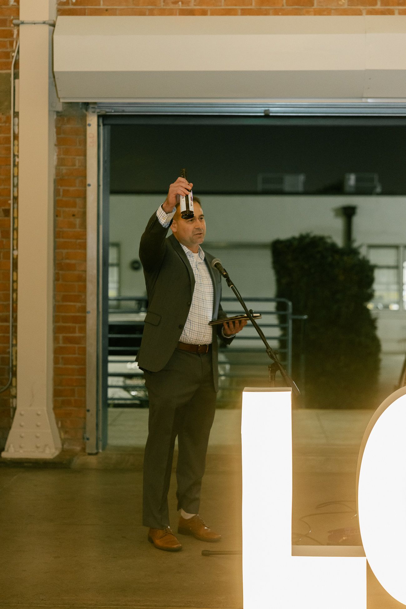 A person in a dark suit raises a glass while speaking into a microphone at an event venue.