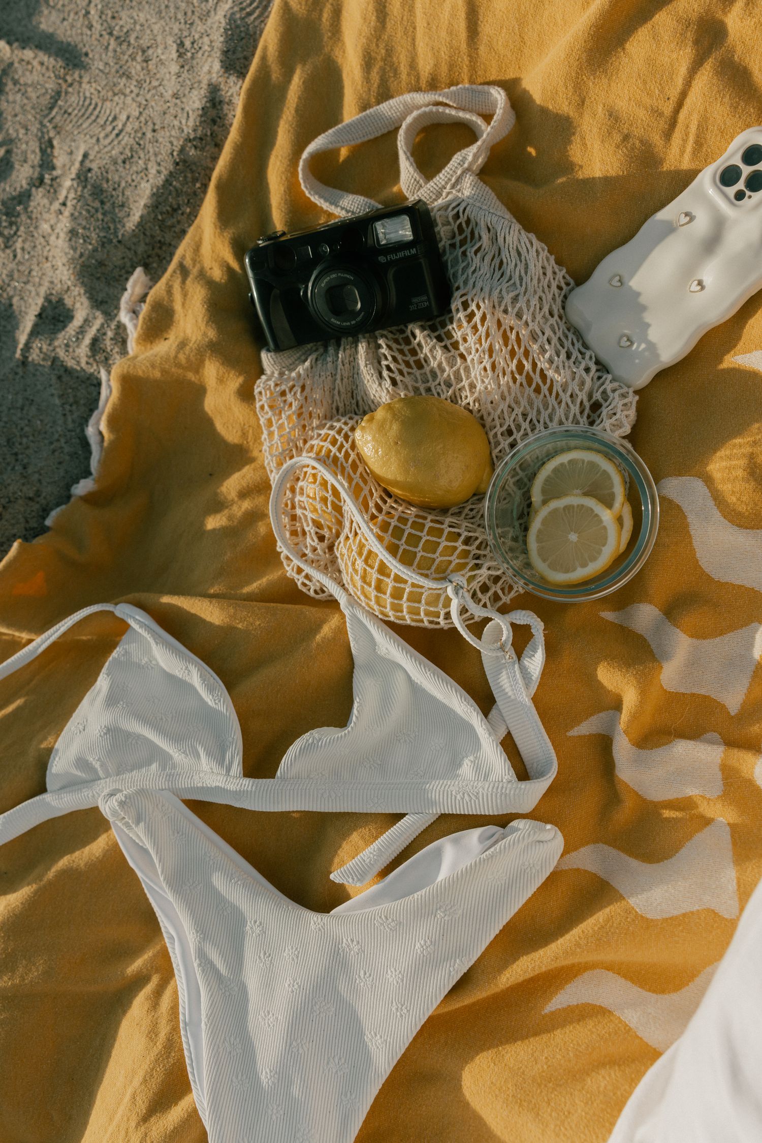Flatlay of beach essentials including white bikini, vintage camera, mesh produce bag with lemons and glass jars on yellow fabric.