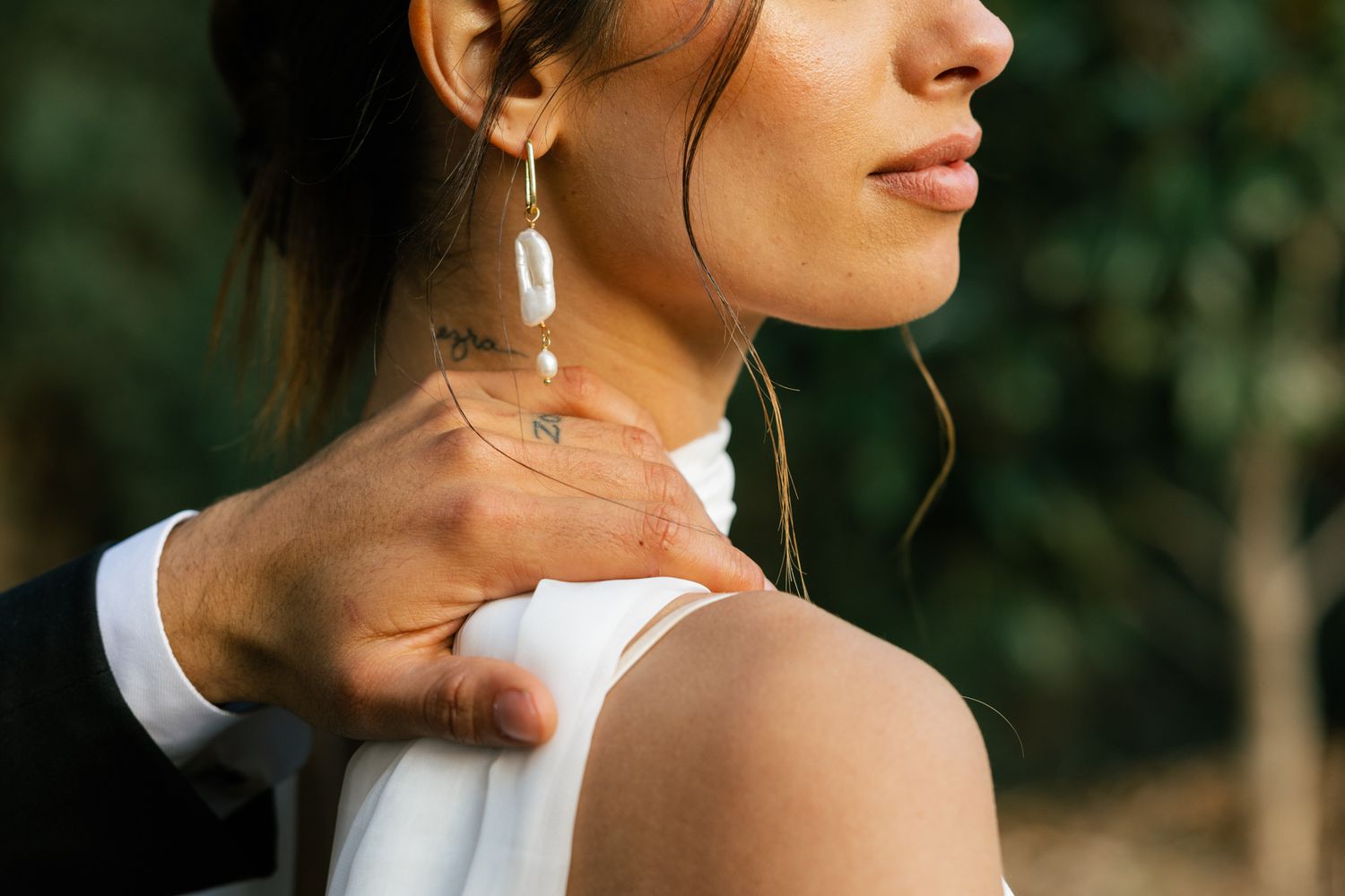 Close-up view of elegant shell earrings and delicate hand touching bare shoulder against blurred natural background.