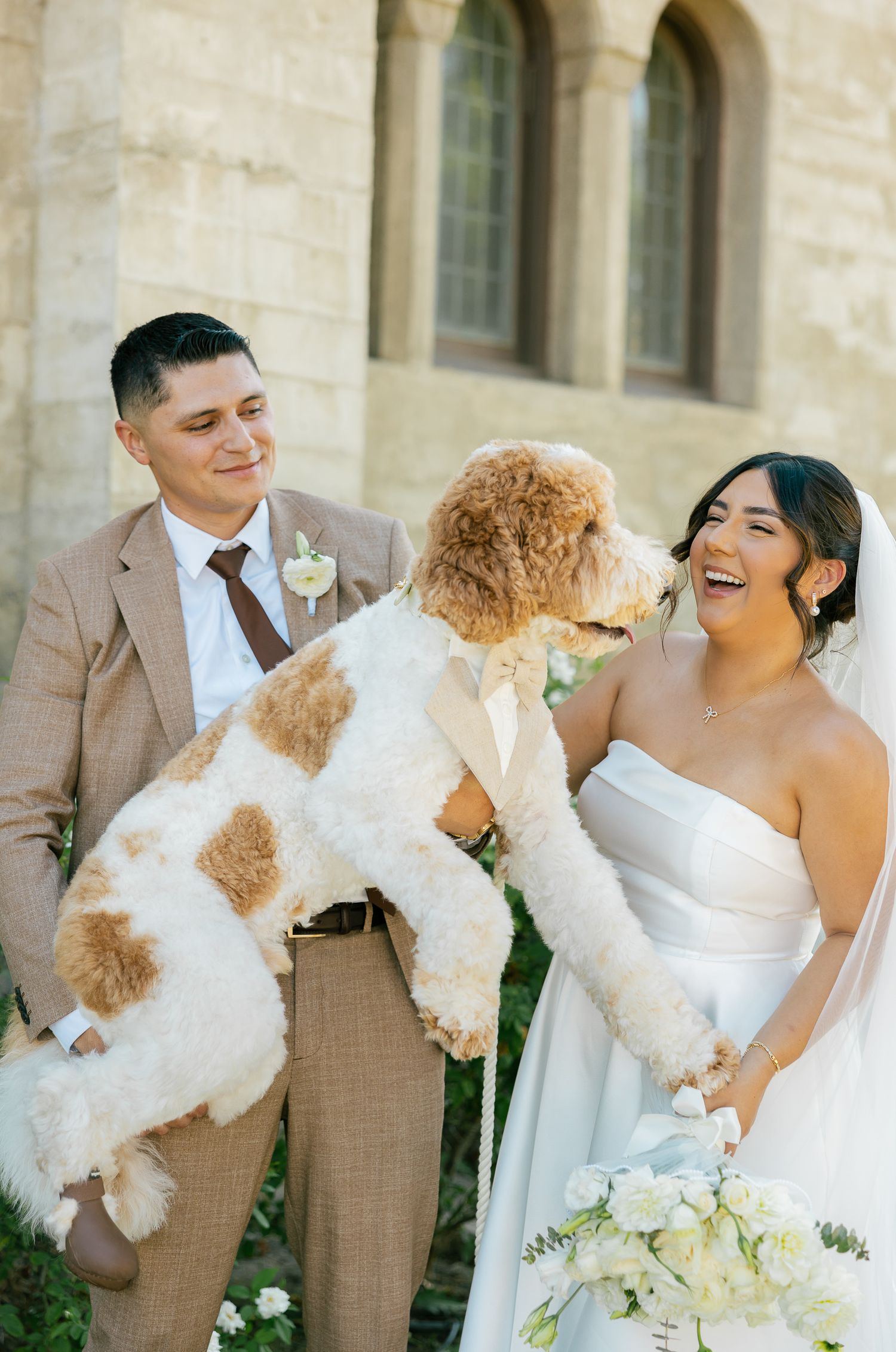 A joyful wedding couple play with their fluffy white and tan dog outside a historic stone building.