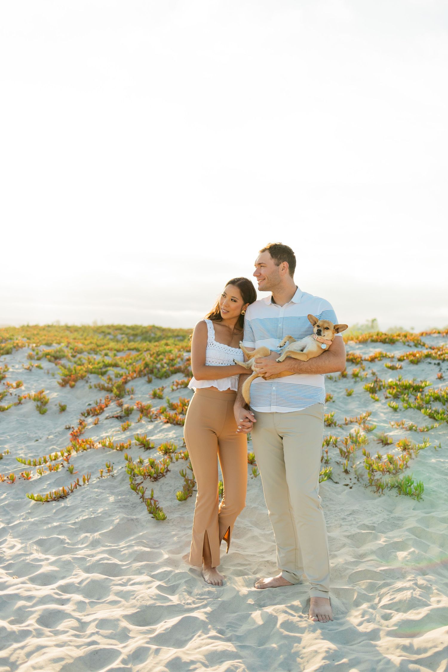 A couple in light colored clothing shares a romantic moment together on a sandy beach with flowering plants.