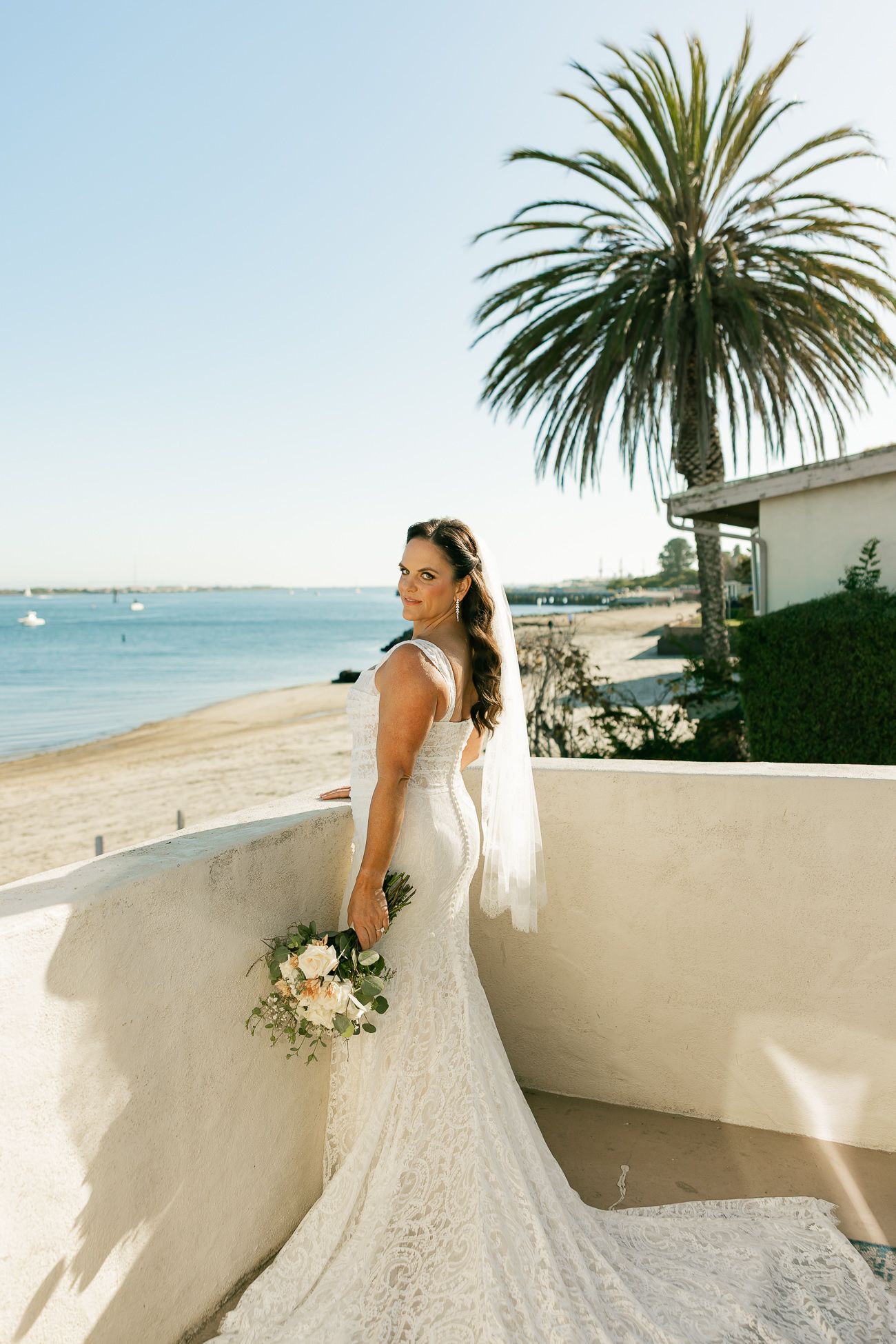 A scenic beach wedding photo with a lace gown, palm tree, and ocean view from a white stucco terrace.