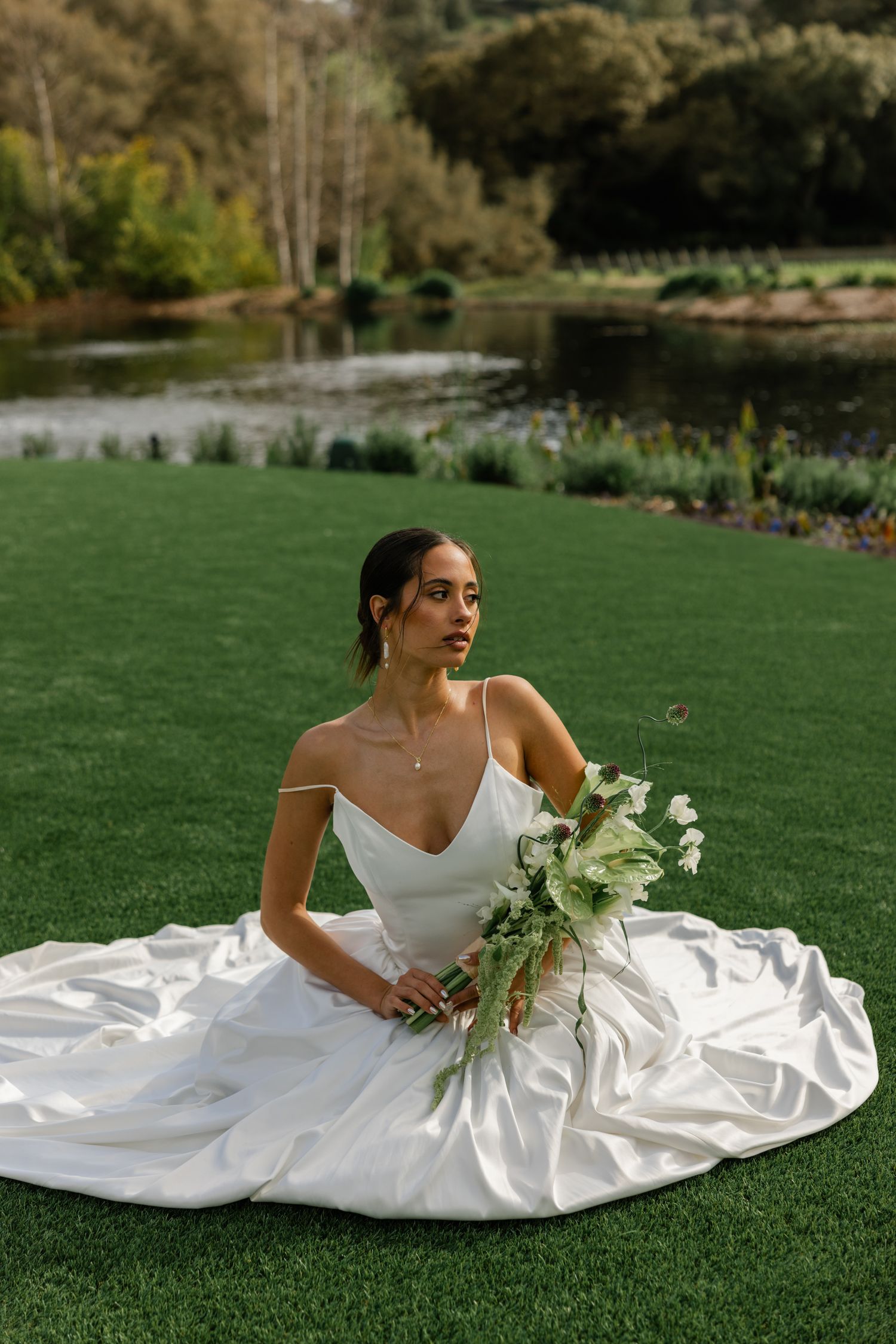 A graceful bride in a white satin gown sits on manicured lawn beside a tranquil pond.