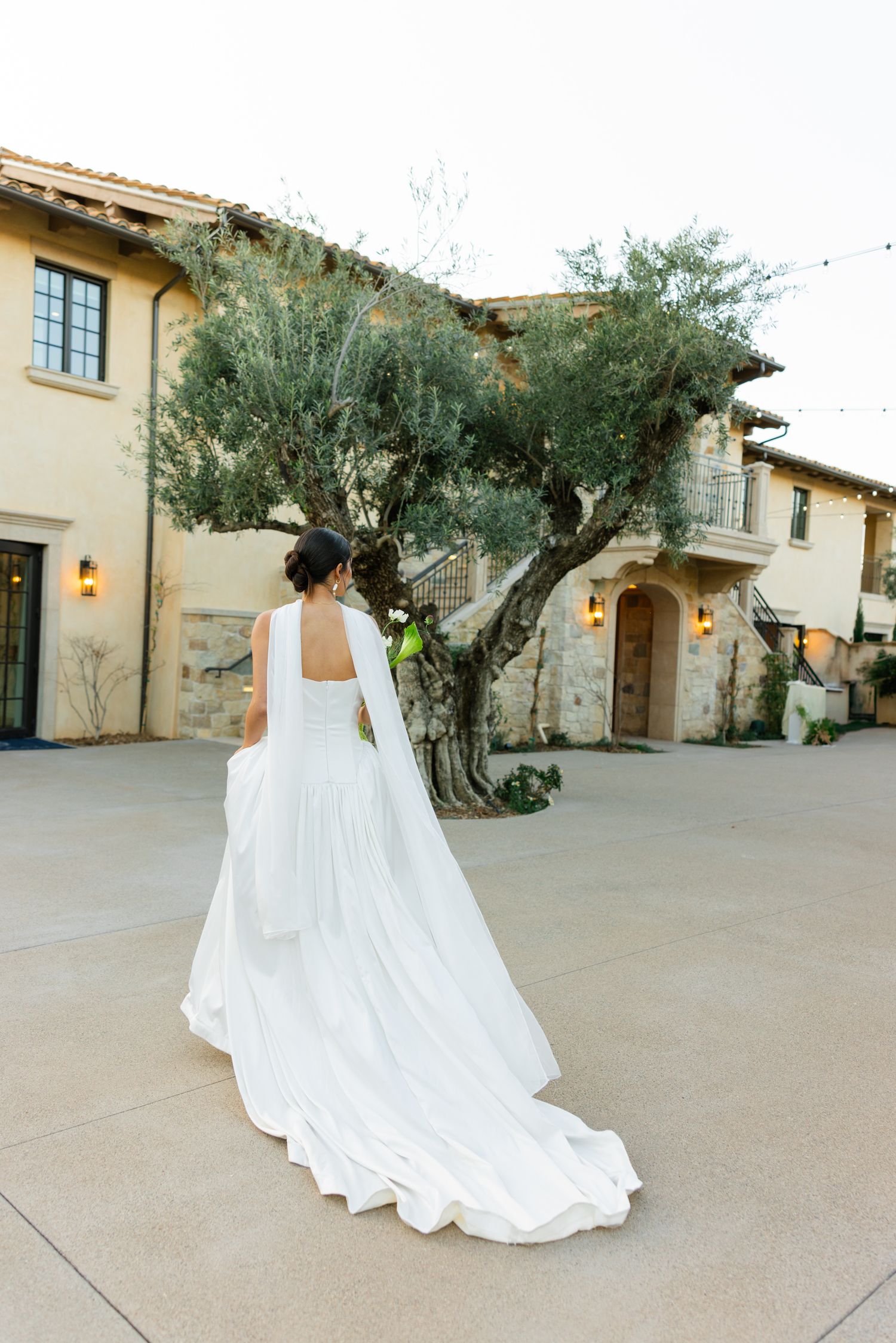 Elegant white wedding dress with flowing train photographed at Mediterranean-style venue with rustic olive tree.