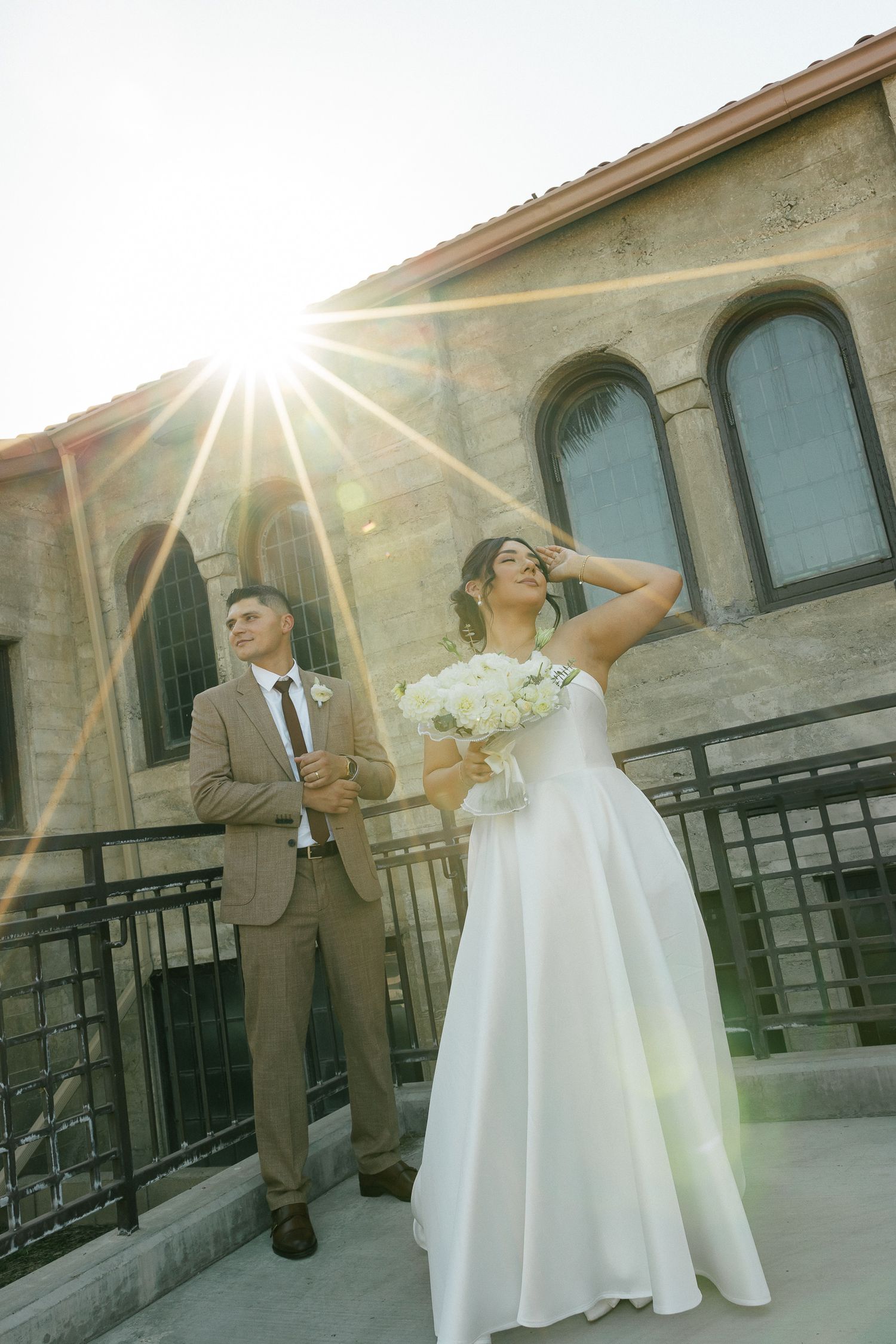 A couple poses on a church balcony during sunset with sunflare backlighting them against arched windows.