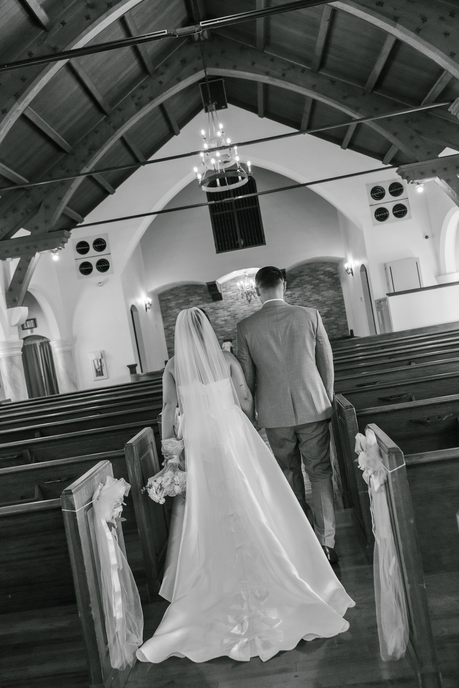 A black and white photo captures a wedding couple walking down a church aisle with vaulted ceilings and wooden pews.