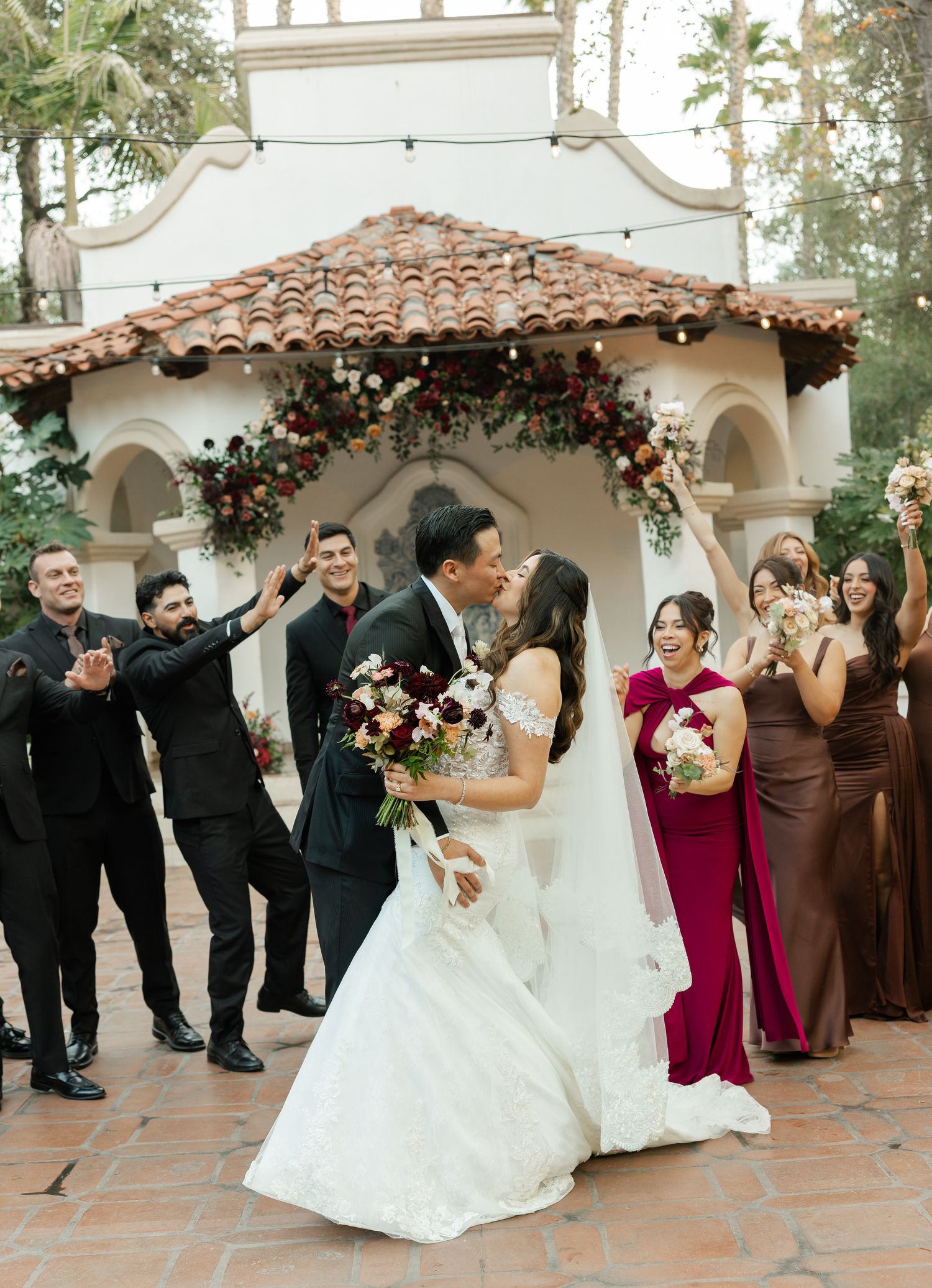 Wedding party poses in a line on Spanish tile courtyard with burgundy and black attire at outdoor venue.