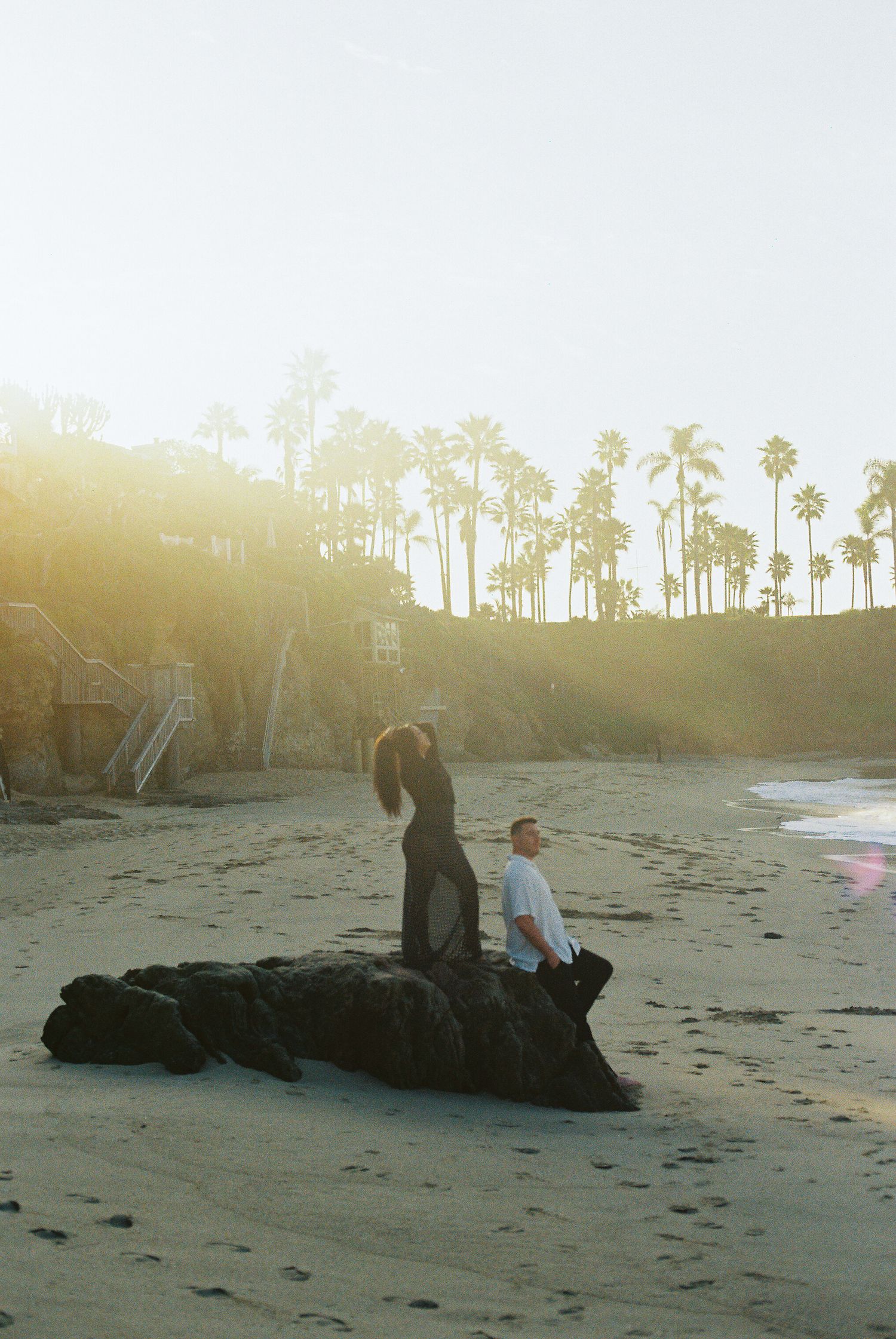 A couple relaxes on a sandy beach at sunset with palm trees silhouetted against the golden sky.