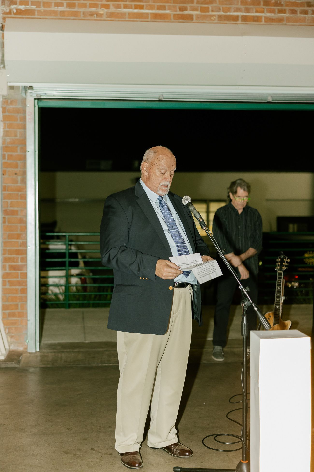 A speaker reads from notes at a podium in an indoor venue with brick walls.