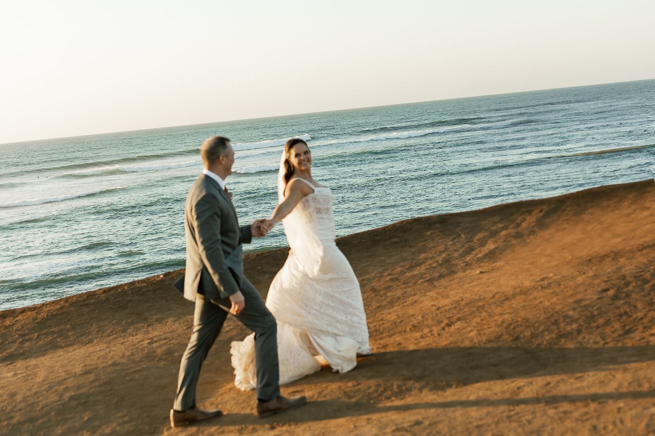 A newlywed couple walks along a scenic coastal cliff at golden hour.
