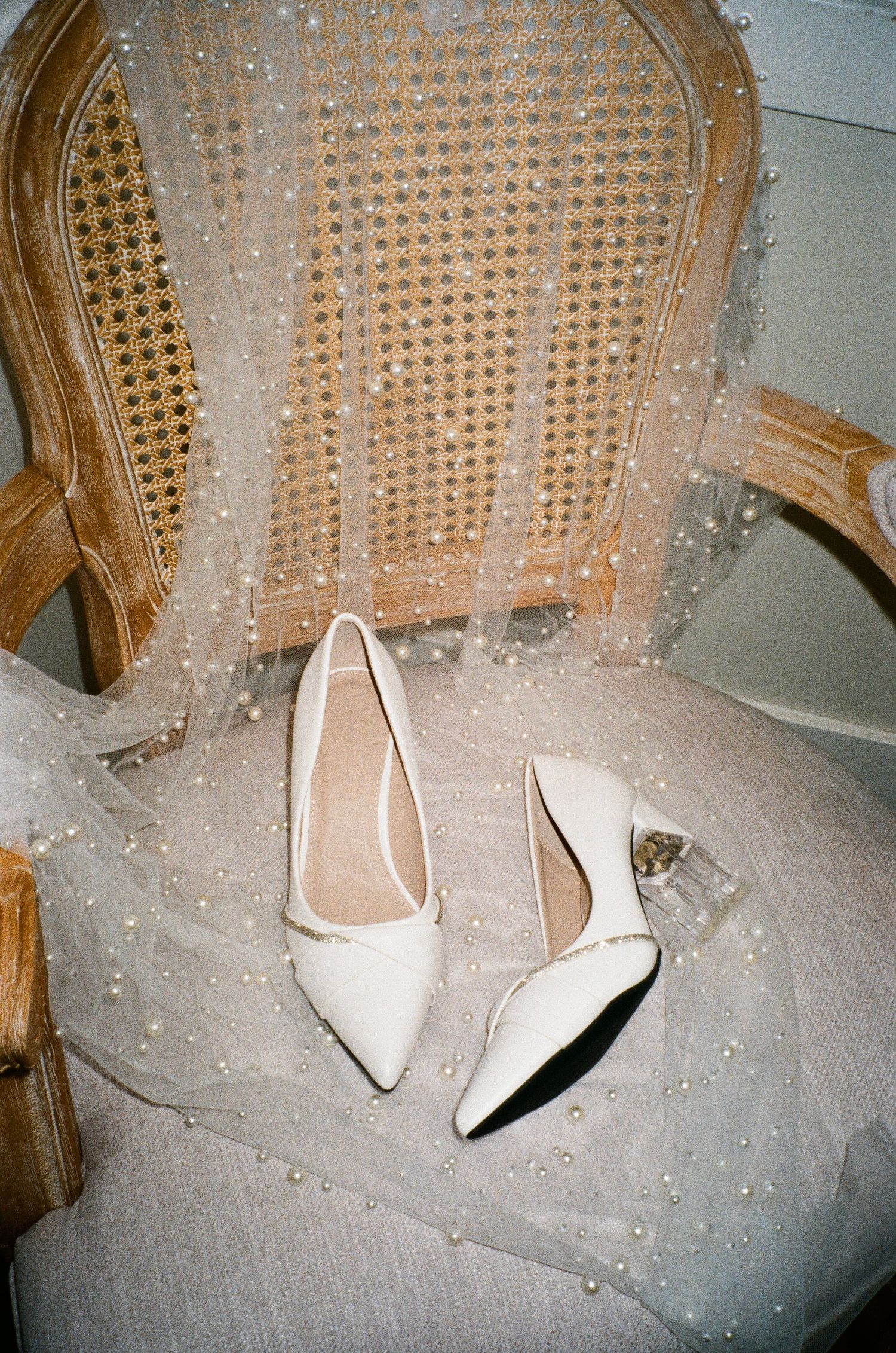 White bridal heels rest elegantly on a delicate tulle veil adorned with scattered pearl embellishments.