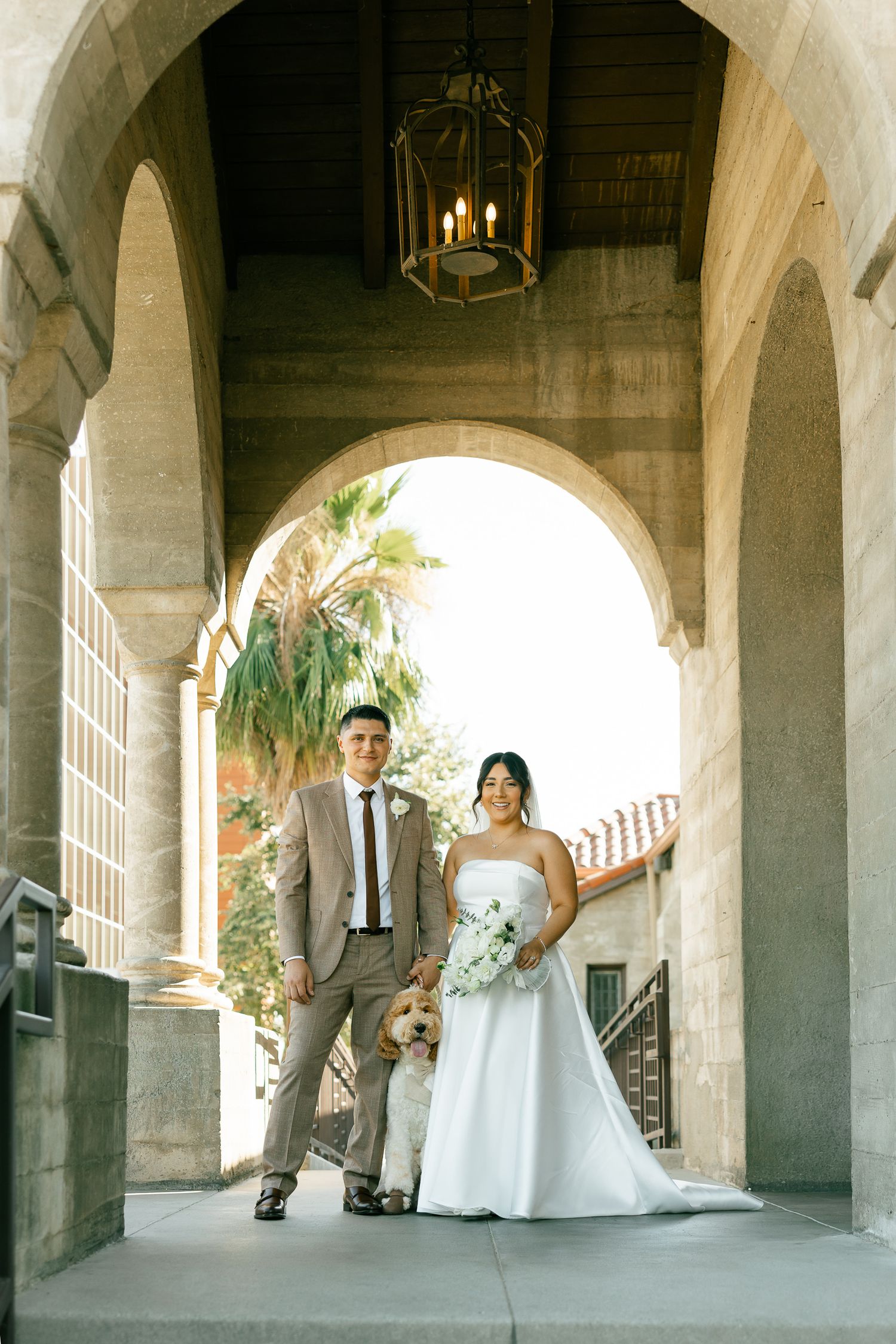 A couple poses underneath stone archways and columns with a hanging chandelier in an elegant architectural setting.